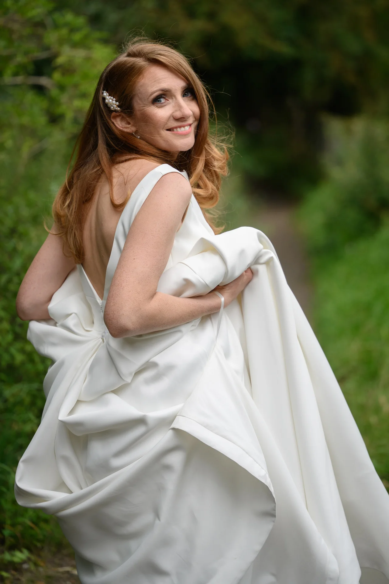 A stunning red-haired bride wearing a satin gown with a v-neckline and open back glances over her shoulder while holding her dress in a lush woodland setting.