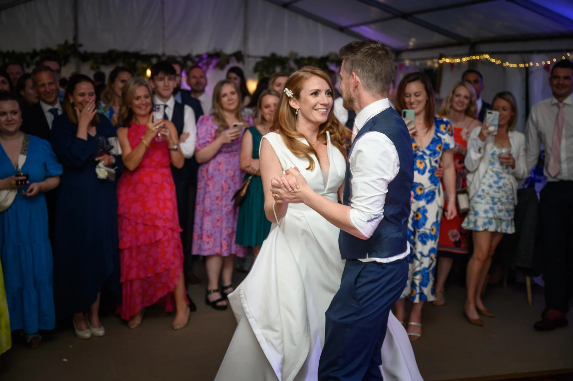 Newlyweds share their first dance together inside a marquee surrounded by cheering and smiling wedding guests.