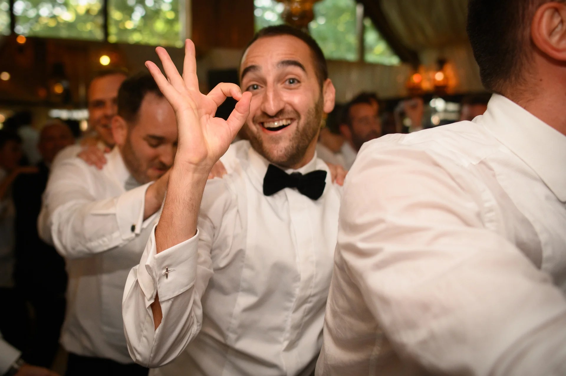 A joyful groom in shirt sleeves and a black bow tie smiles and gestures with an okay sign while leading a conga line at an evening wedding reception.
