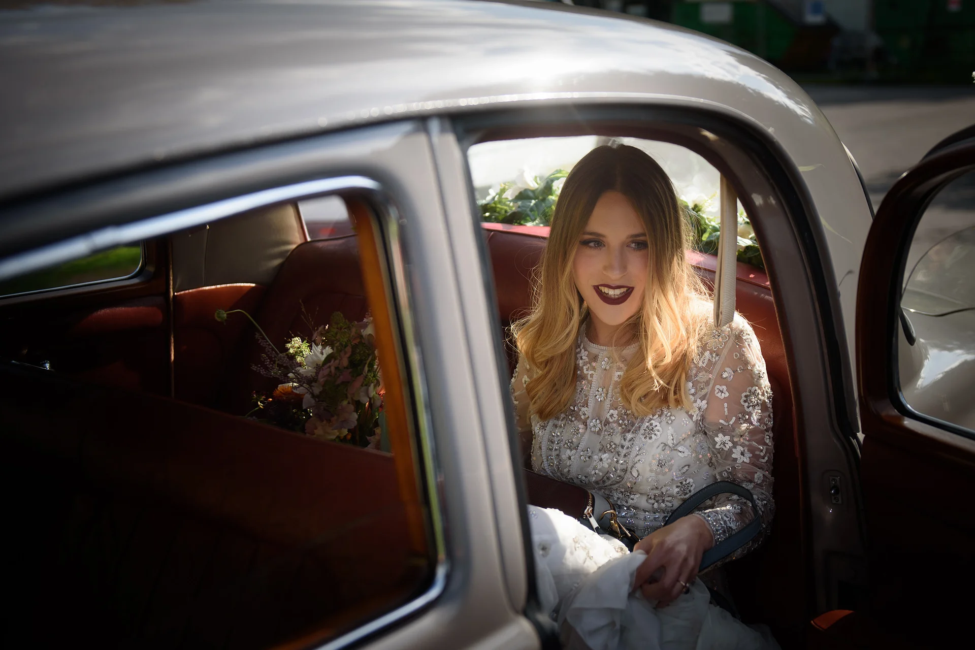 A smiling bride with long blonde hair and an embellished long-sleeve gown sits inside the leather interior of a classic vintage wedding car.