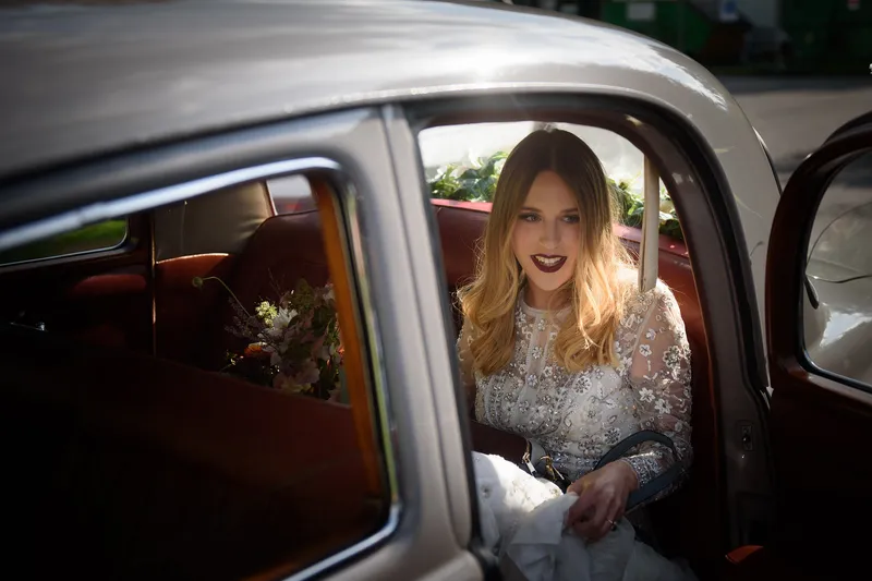 A smiling bride with long blonde hair and an embellished long-sleeve gown sits inside the leather interior of a classic vintage wedding car.