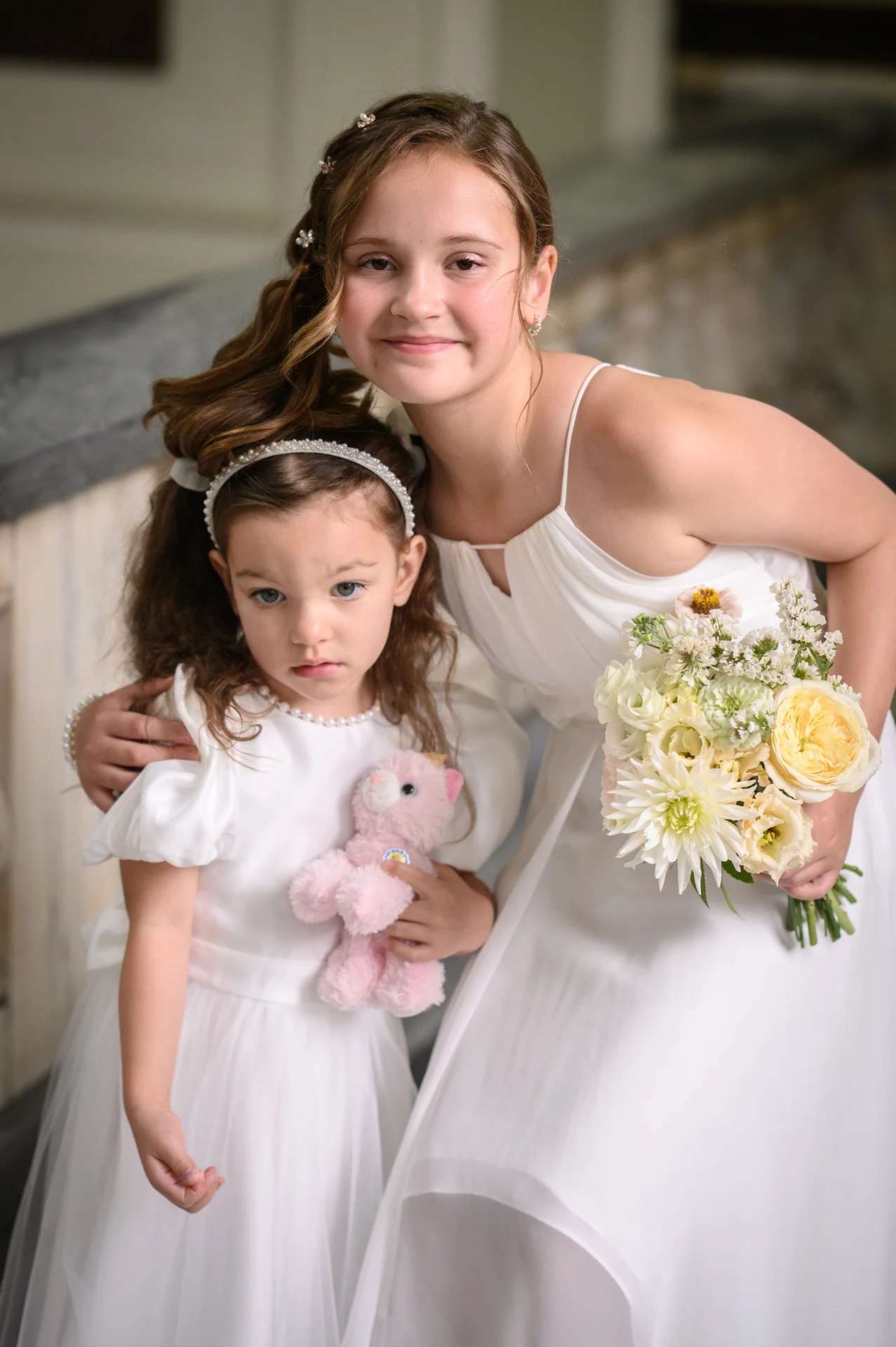 An older bridesmaid in a white gown holding a bouquet stands behind a younger flower girl wearing a white dress and headband while holding a pink toy.