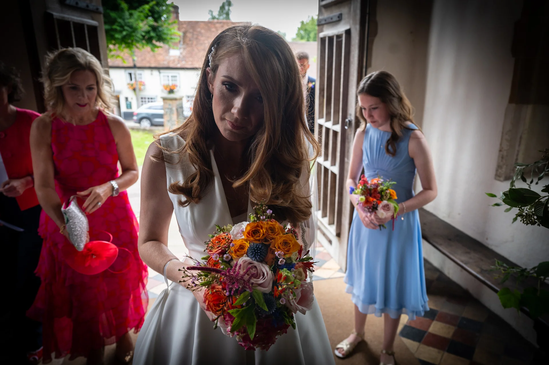 A bride with long brown hair looks down at her colourful autumn bouquet inside a church doorway, surrounded by her bridesmaids and family members before the ceremony.