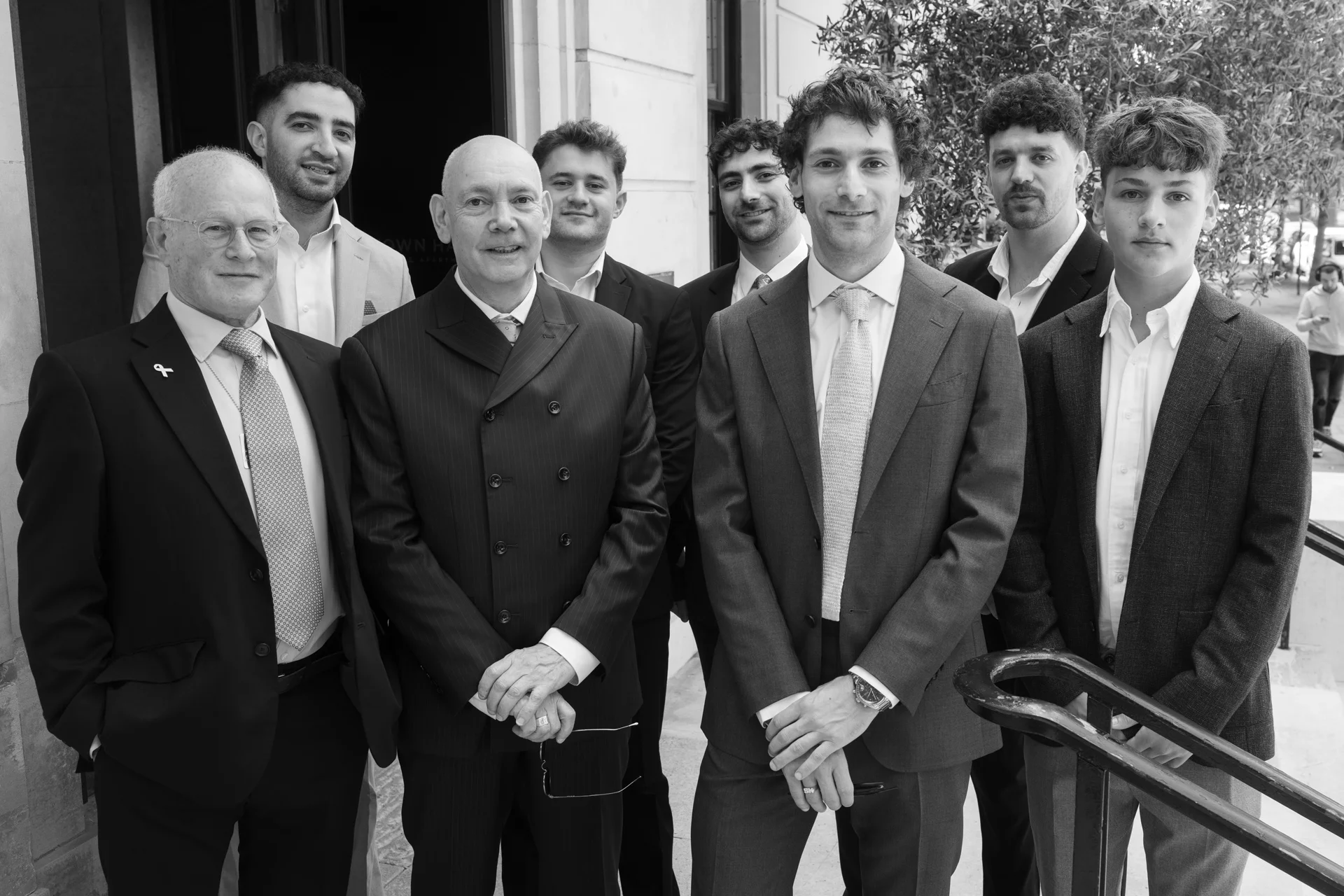 A sophisticated black and white group portrait of the groom and his groomsmen posing together outside a city venue wearing tailored suits and ties.