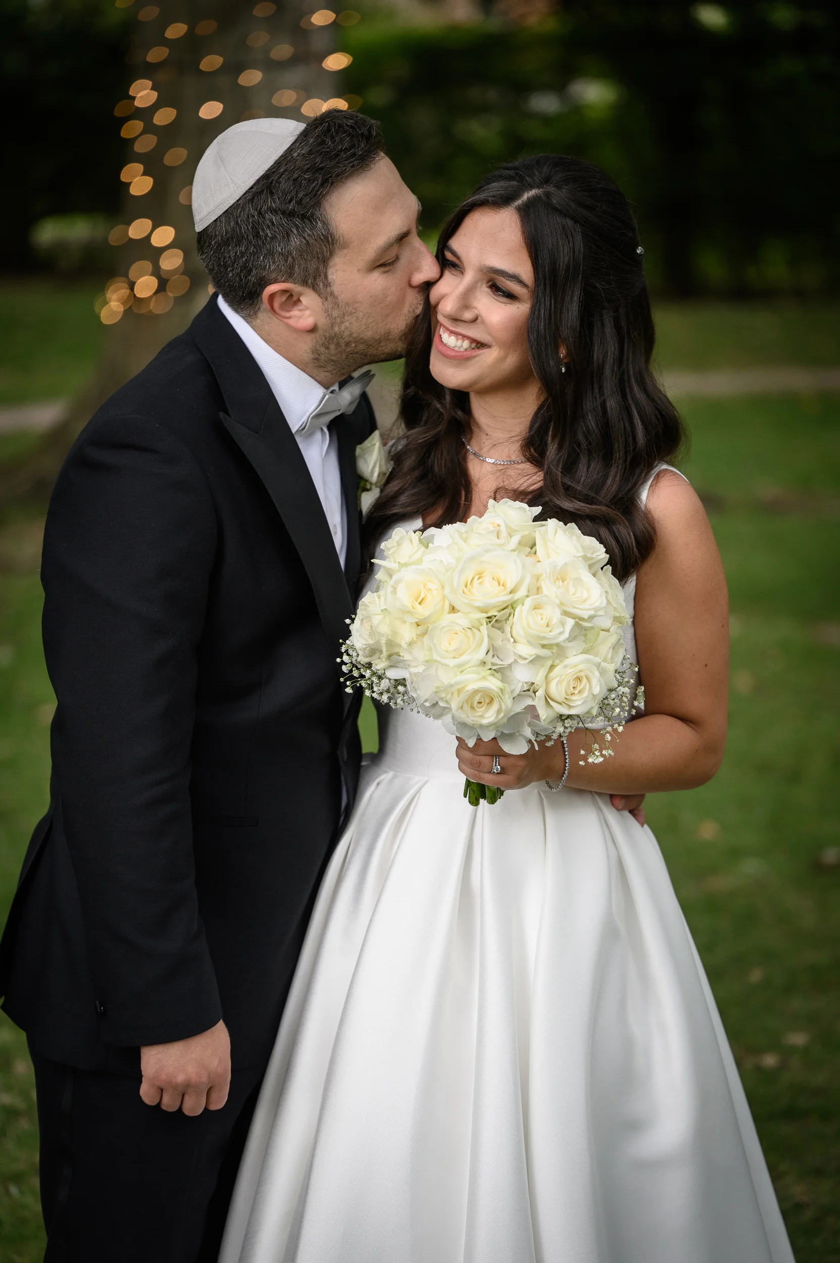 A groom wearing a kippah and black tuxedo kisses his smiling bride on the cheek as she holds a round bouquet of white roses outdoors.