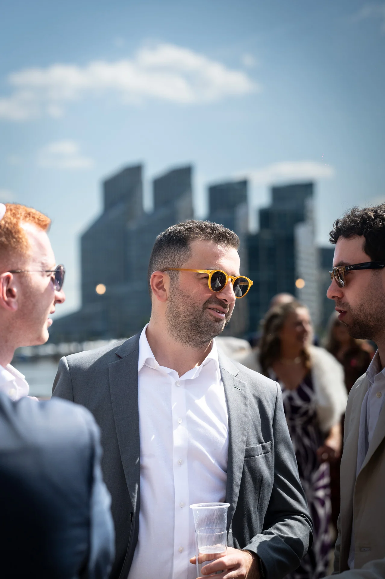 Several wedding guests mingle outdoors during a daytime drinks reception, with a modern city skyline visible in the background under a blue sky.