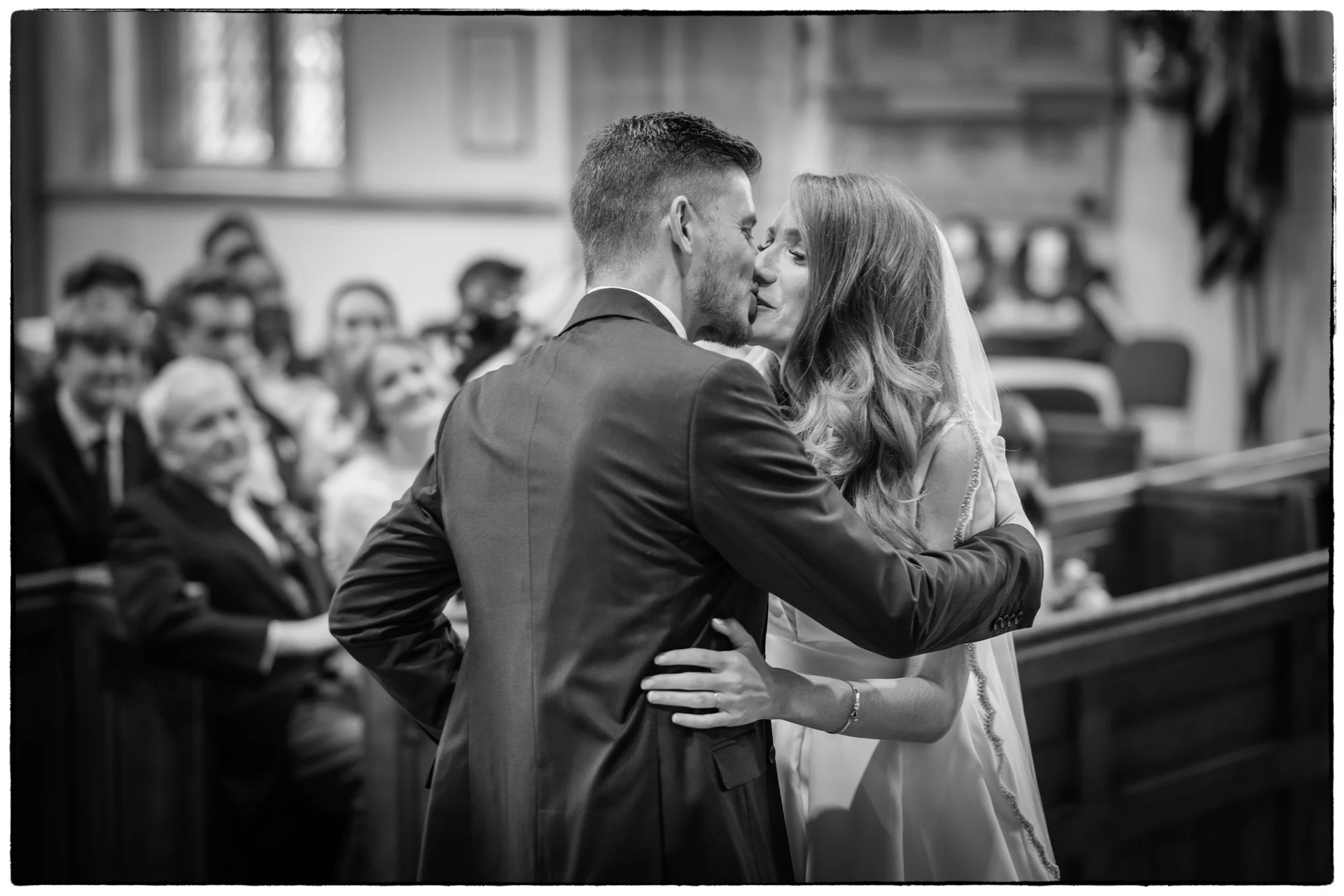 The newly married couple share their first kiss at the altar during a traditional church wedding ceremony, with seated guests watching on in the background.
