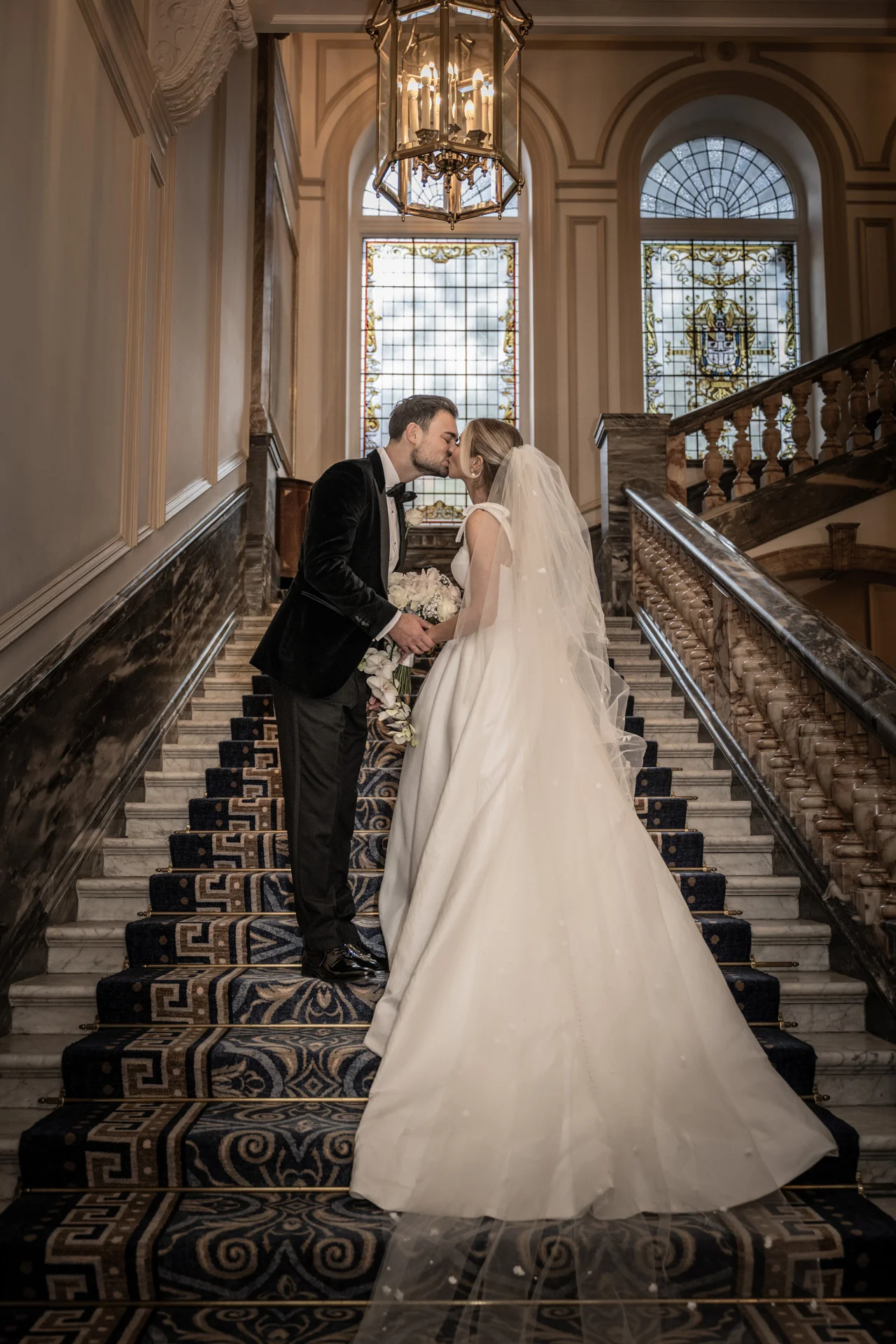 A bride and groom share a tender kiss while standing on a grand carpeted staircase beneath stained glass windows and a classic chandelier.