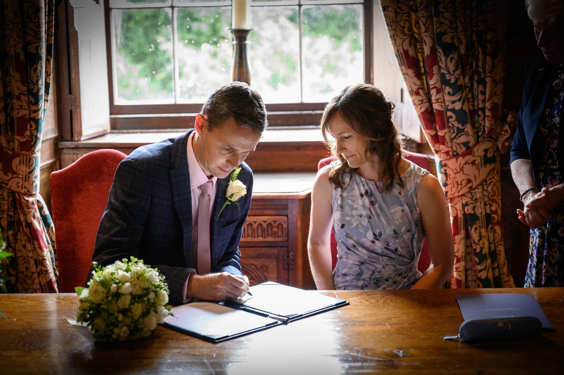 A groom in a checked suit signs the marriage register while his bride looks on lovingly beside him in a room with patterned curtains and natural window light.