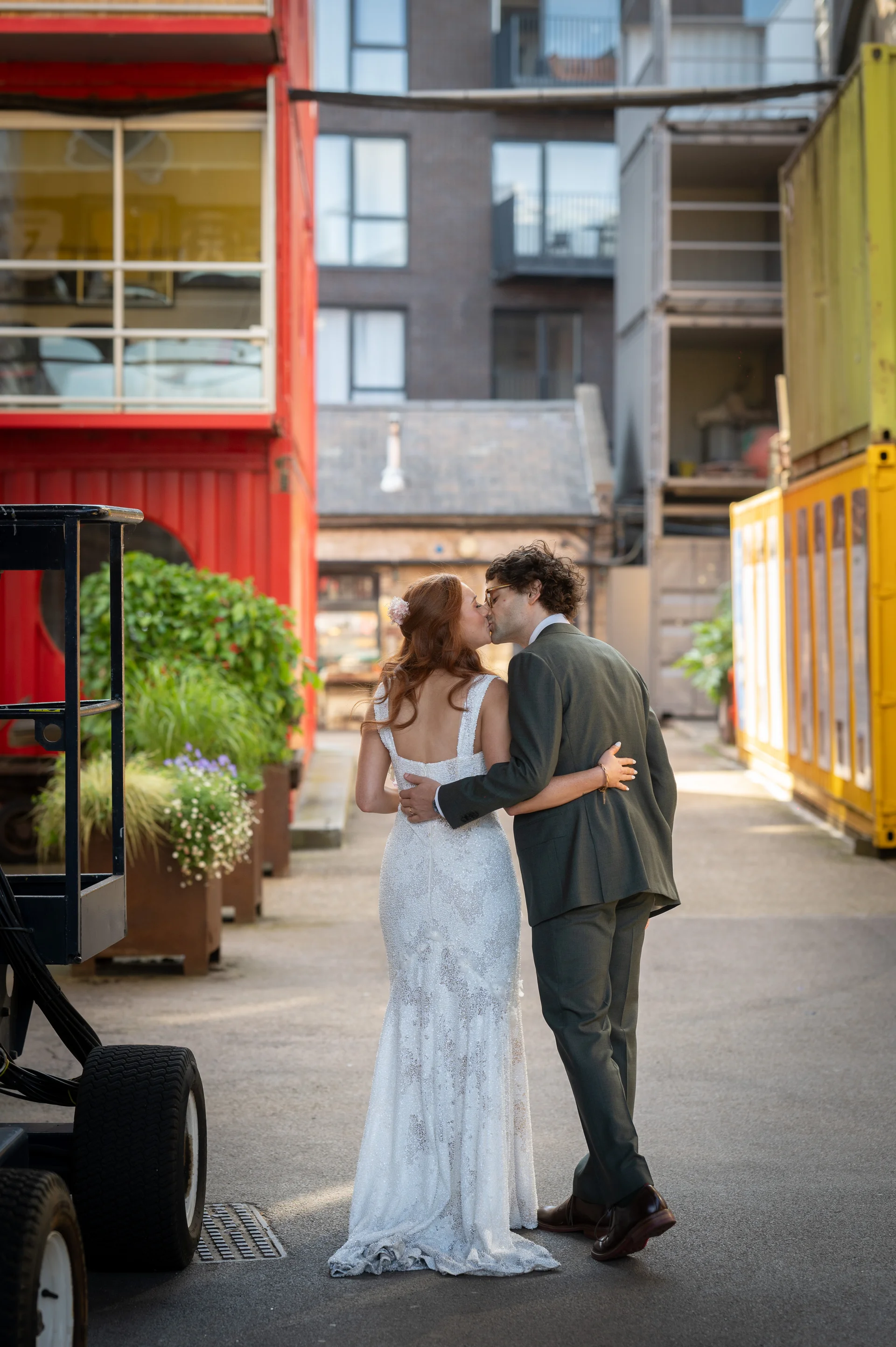 A newlywed couple embraces and kisses in an industrial urban alleyway framed by colourful shipping containers and modern architecture.