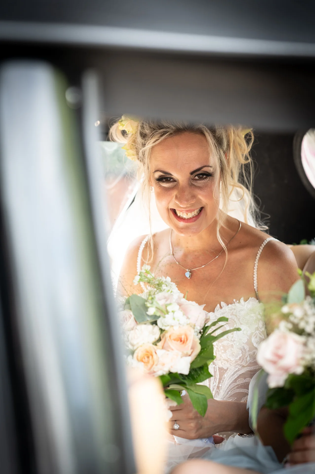 A smiling bride holding a floral bouquet looks out through the window of a wedding car while wearing a lace gown and delicate necklace.