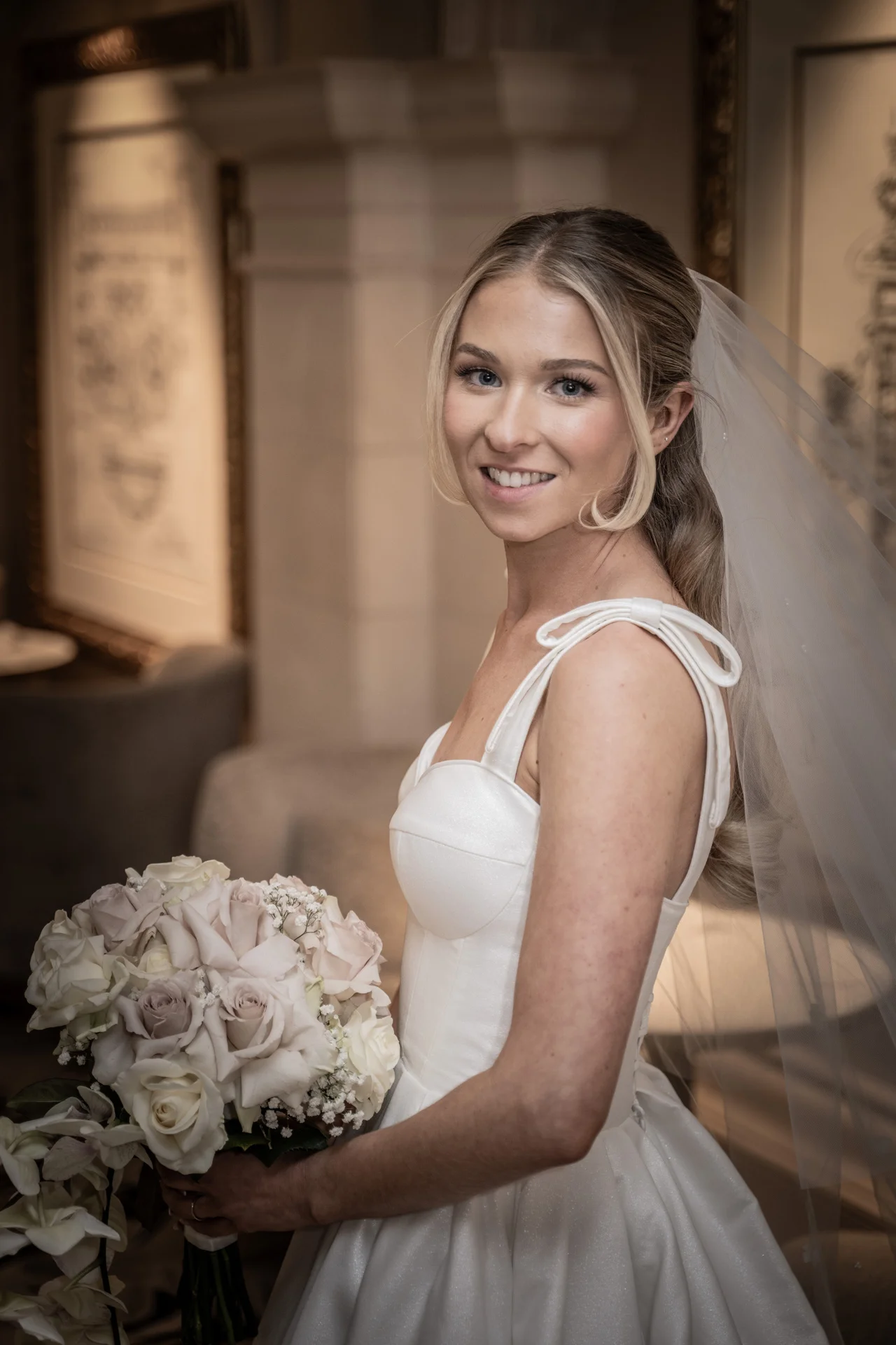 A smiling bride wearing a structured bodice gown and veil holds a bouquet of roses and baby's breath indoors.