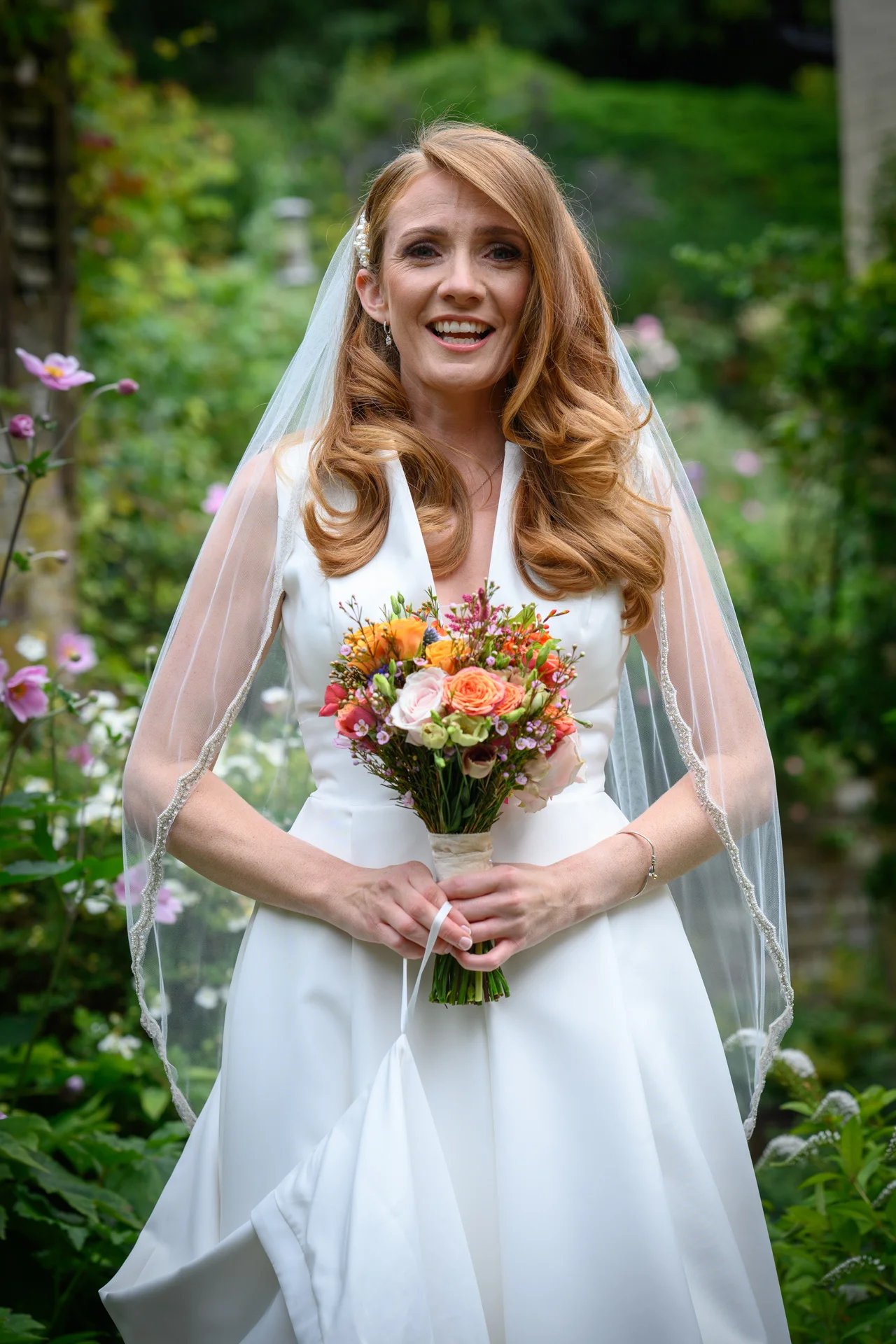 A smiling bride with long red hair and a white satin v-neck gown holds a colourful hand-tied bouquet in a lush garden.