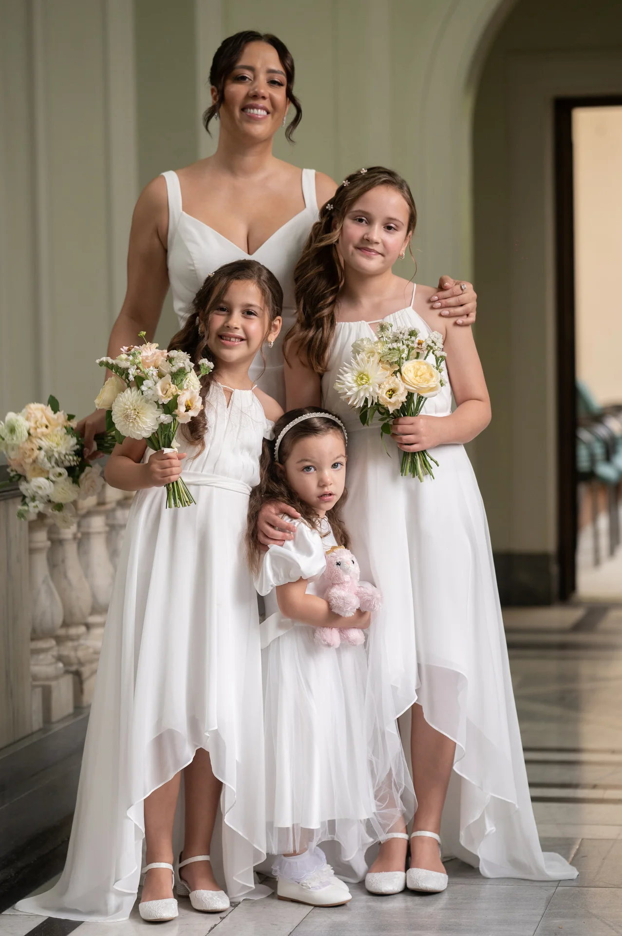 An adult bridesmaid and three young flower girls in matching white dresses pose indoors holding bouquets of cream and white flowers.