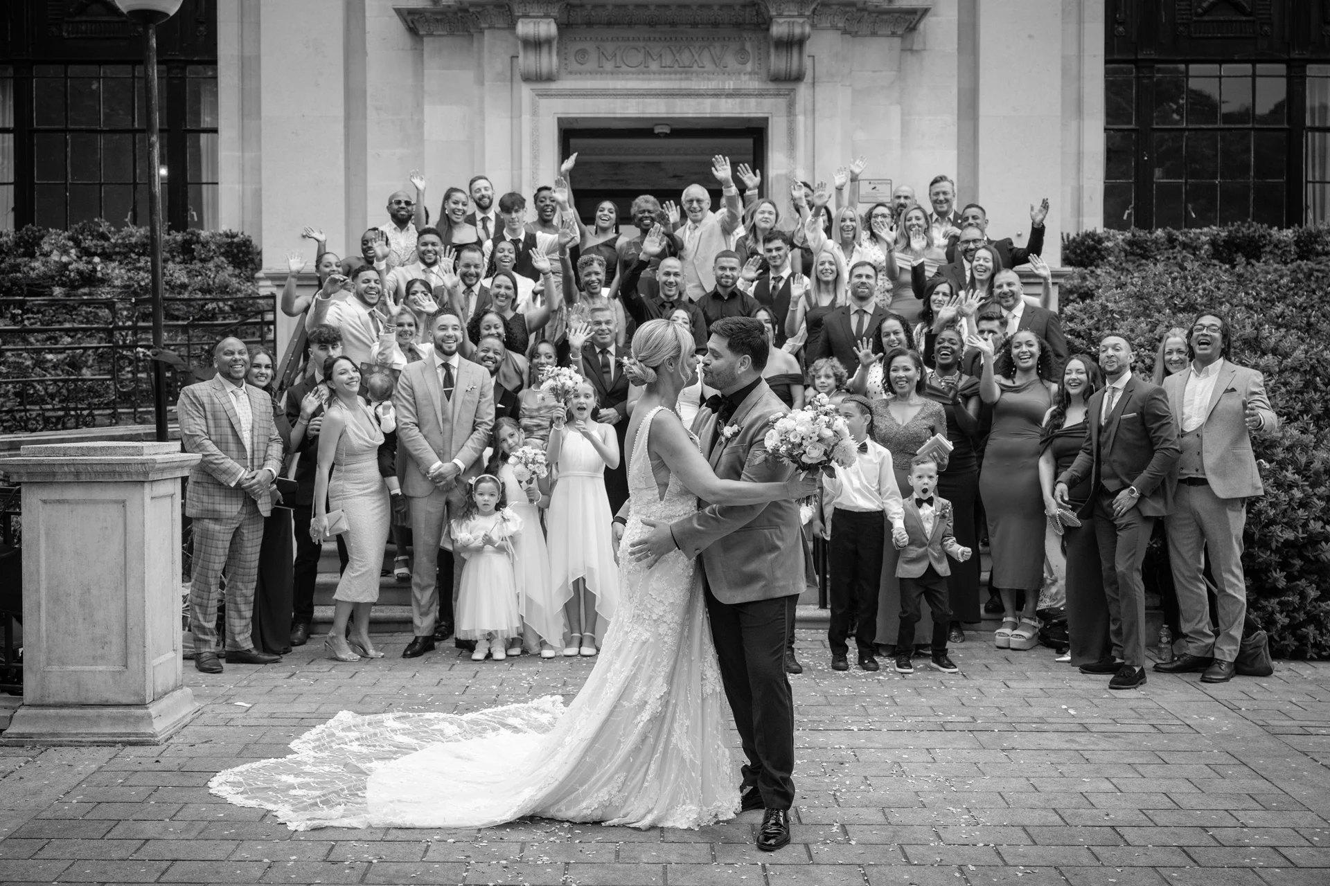 A newlywed couple embraces and kisses in the courtyard of a stately building while a large crowd of cheering guests waves from the steps behind them.