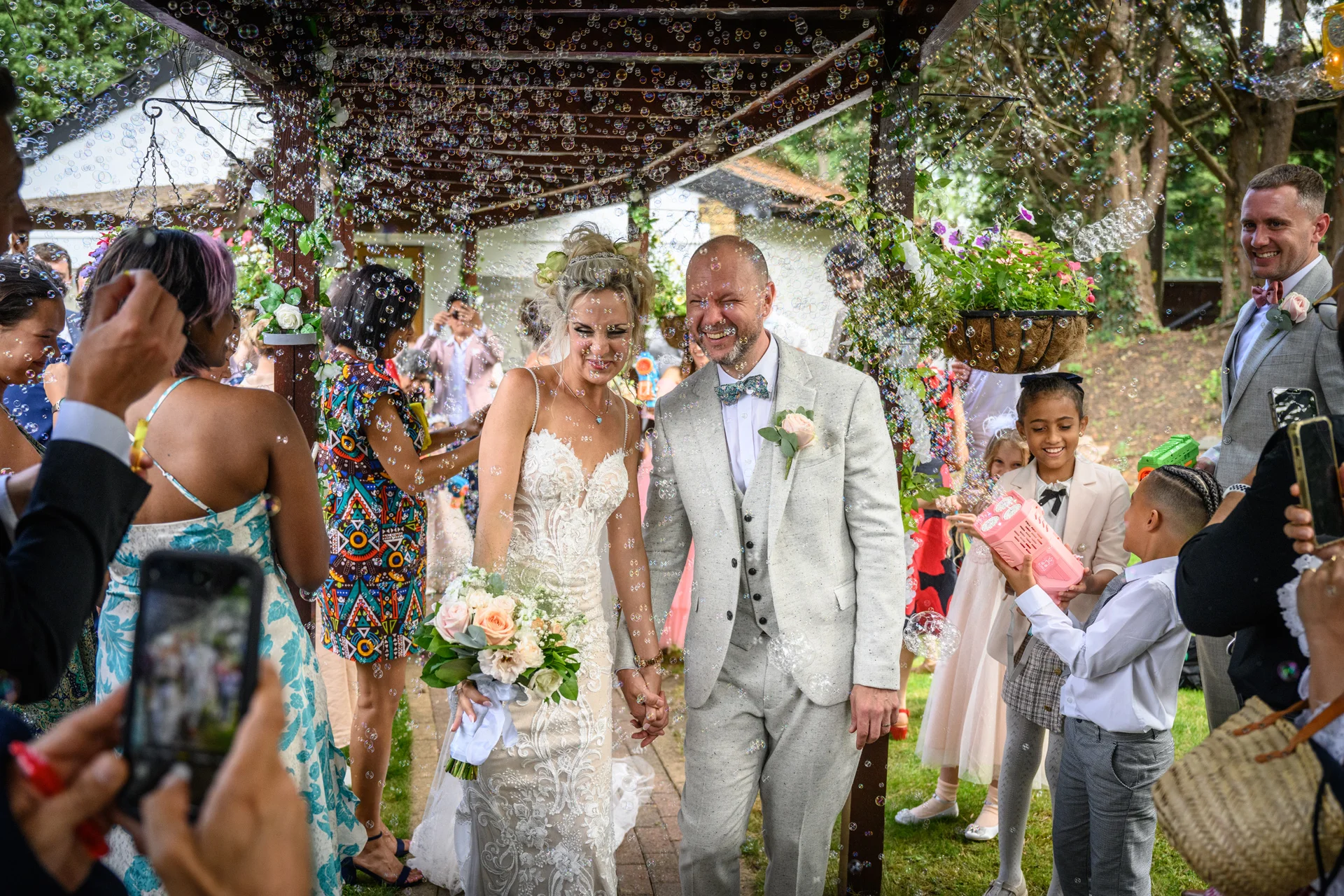 A newlywed couple walks hand in hand beneath a wooden pergola as cheering guests blow bubbles around them, with the bride holding a romantic pastel bouquet.