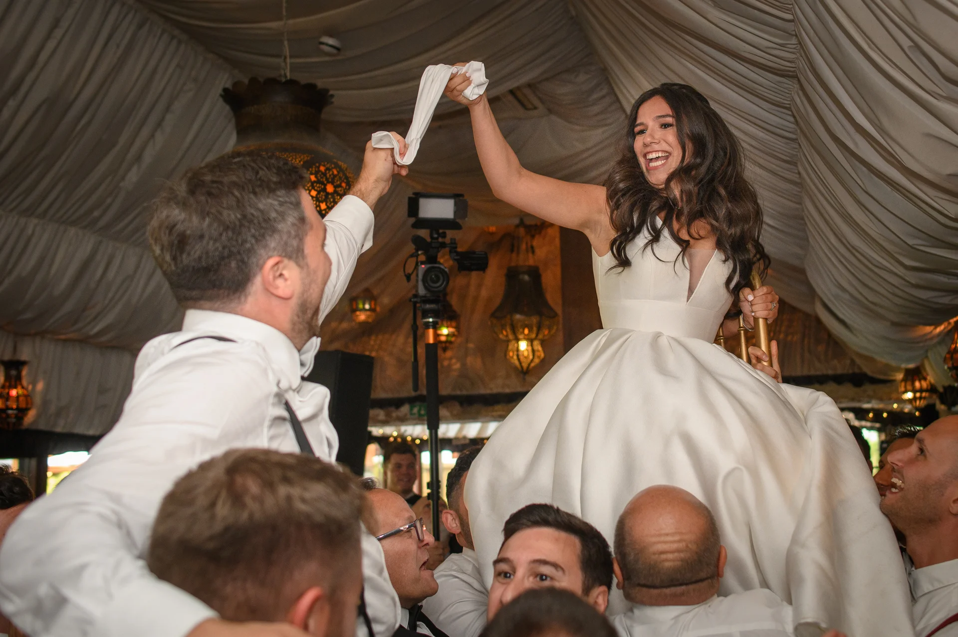 A joyful bride wearing a satin A-line gown is lifted high in the air on a chair during the lively hora chair lift at a wedding reception inside a draped marquee.