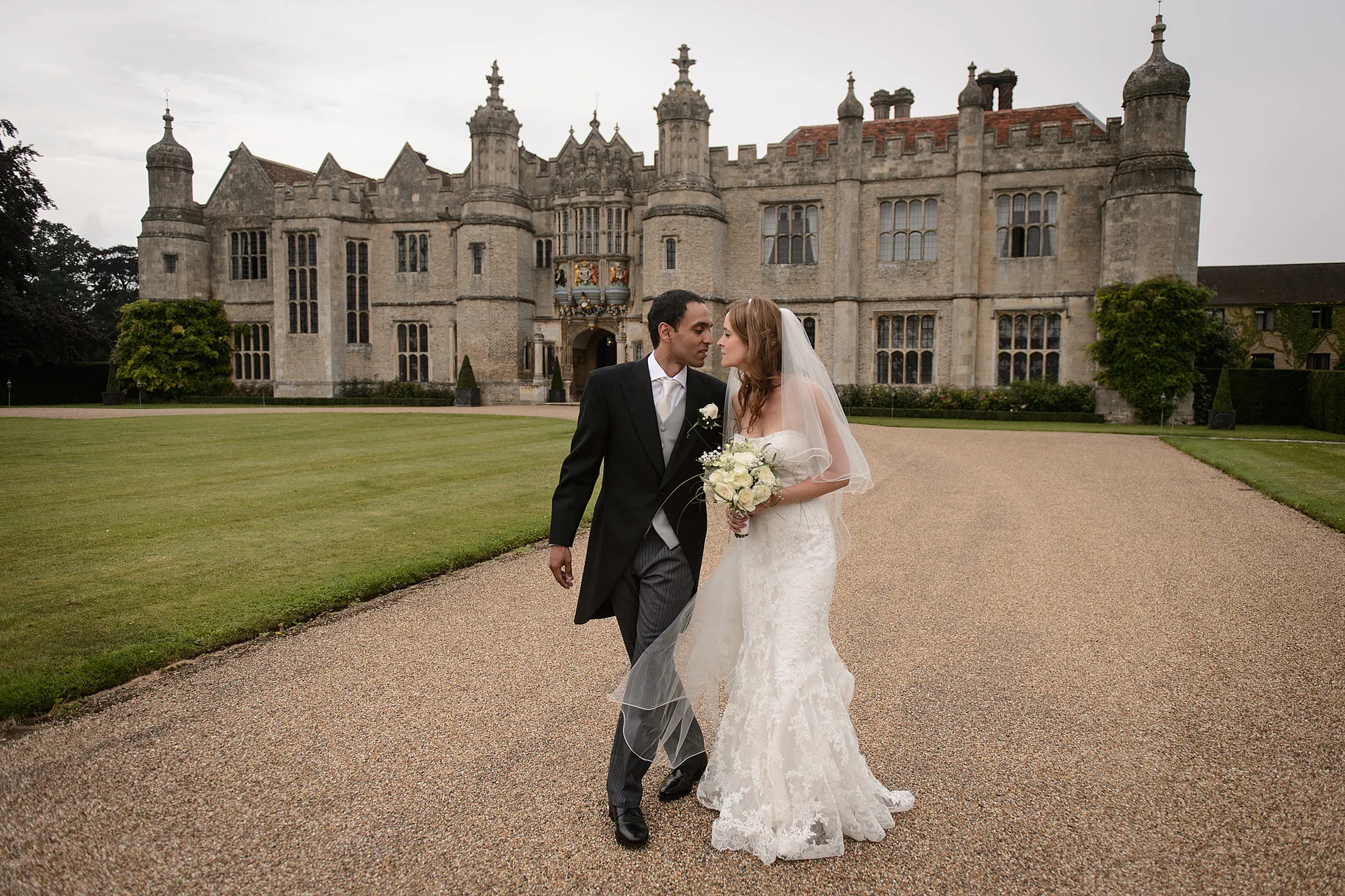 A newlywed couple walks hand in hand along a gravel driveway in front of a grand stone country house, with the bride holding a white bouquet and wearing a lace gown.