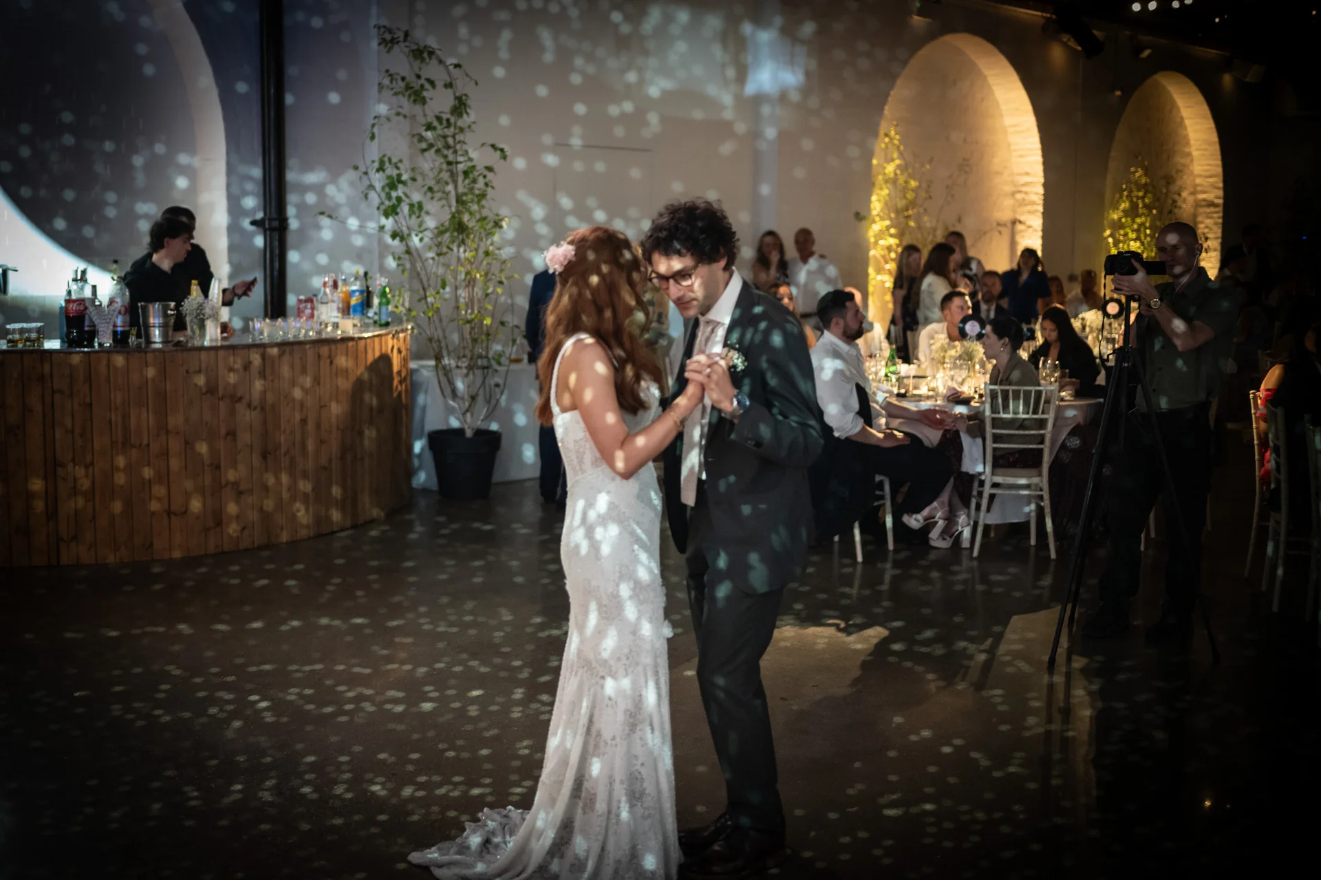 A newlywed couple shares their first dance in a spacious industrial warehouse venue lit with speckled projection lighting while guests watch from tables.