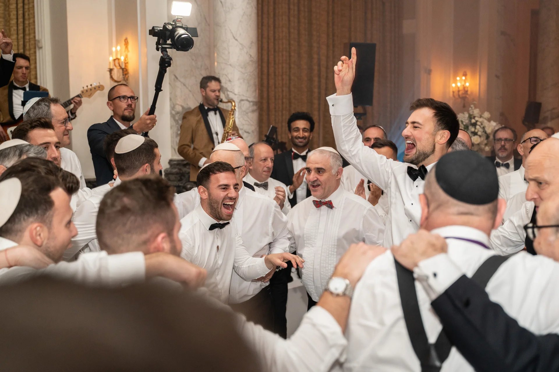 A joyful groom and wedding guests in white shirts and black bow ties celebrate enthusiastically on a crowded dance floor during the evening reception.