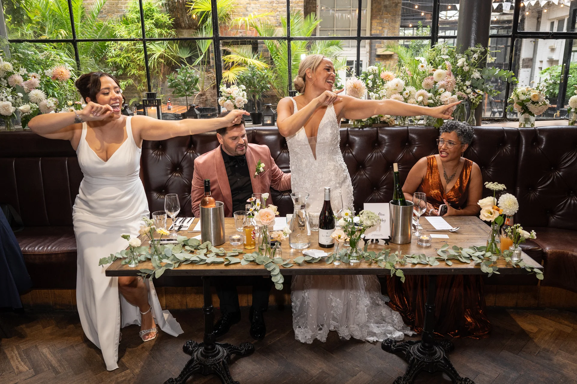 Two brides and wedding guests laugh and dance excitedly behind a wooden banquet table adorned with greenery runners and wine bottles in a rustic industrial restaurant venue.