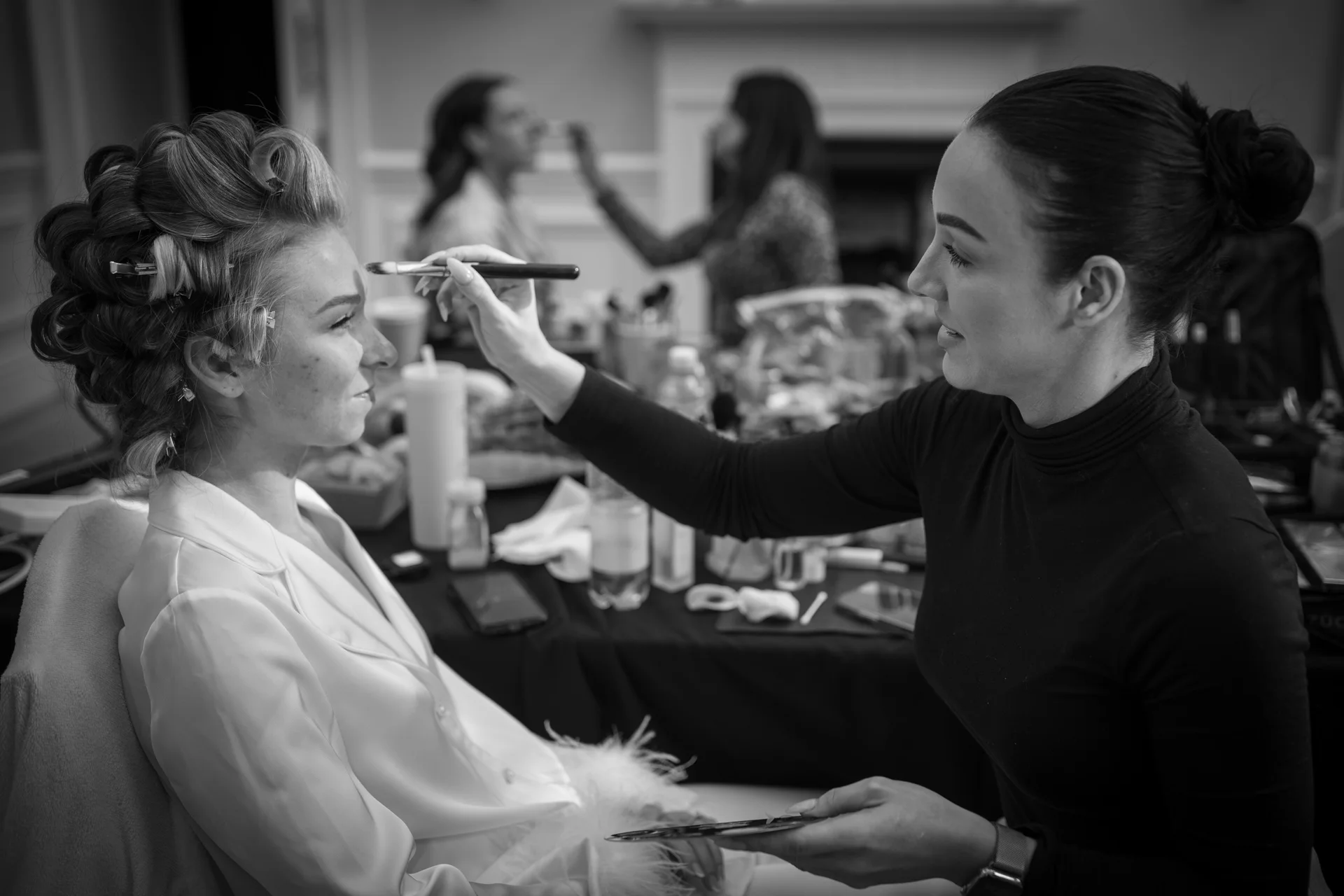 A black and white photograph captures a bride having her makeup applied by a professional artist while rollers are still set in her hair during bridal preparations.