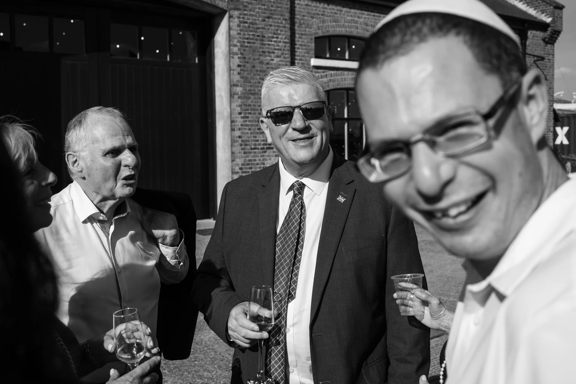 A candid black and white shot of wedding guests mingling outside during a drinks reception, featuring a smiling man in the foreground wearing a kippah and glasses.
