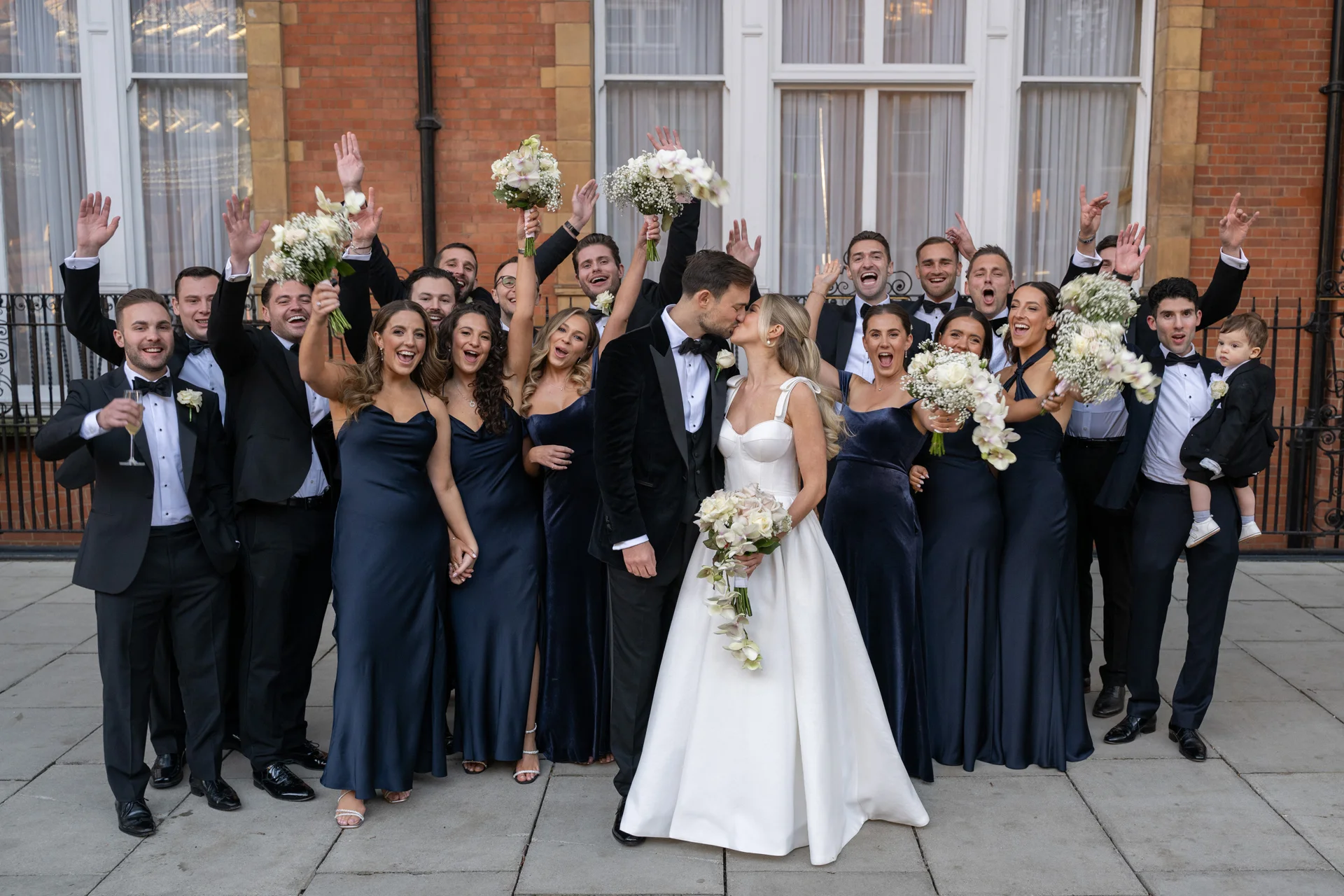 The bride and groom share a kiss in front of their large wedding party, who raise their hands and bouquets in celebration outside a historic brick building.