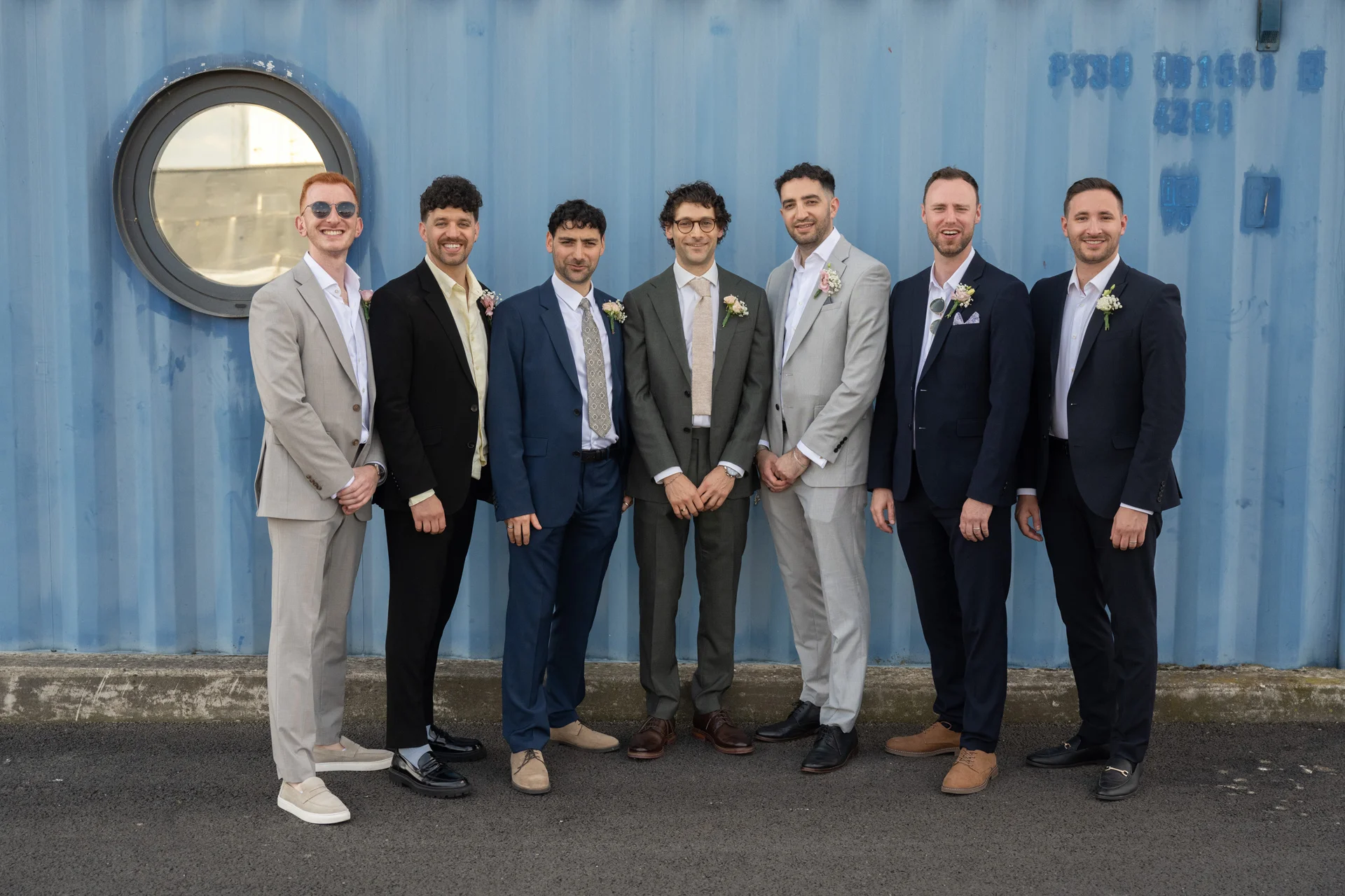 A group of seven smiling groomsmen in mismatched suits pose together outside against a textured blue shipping container.