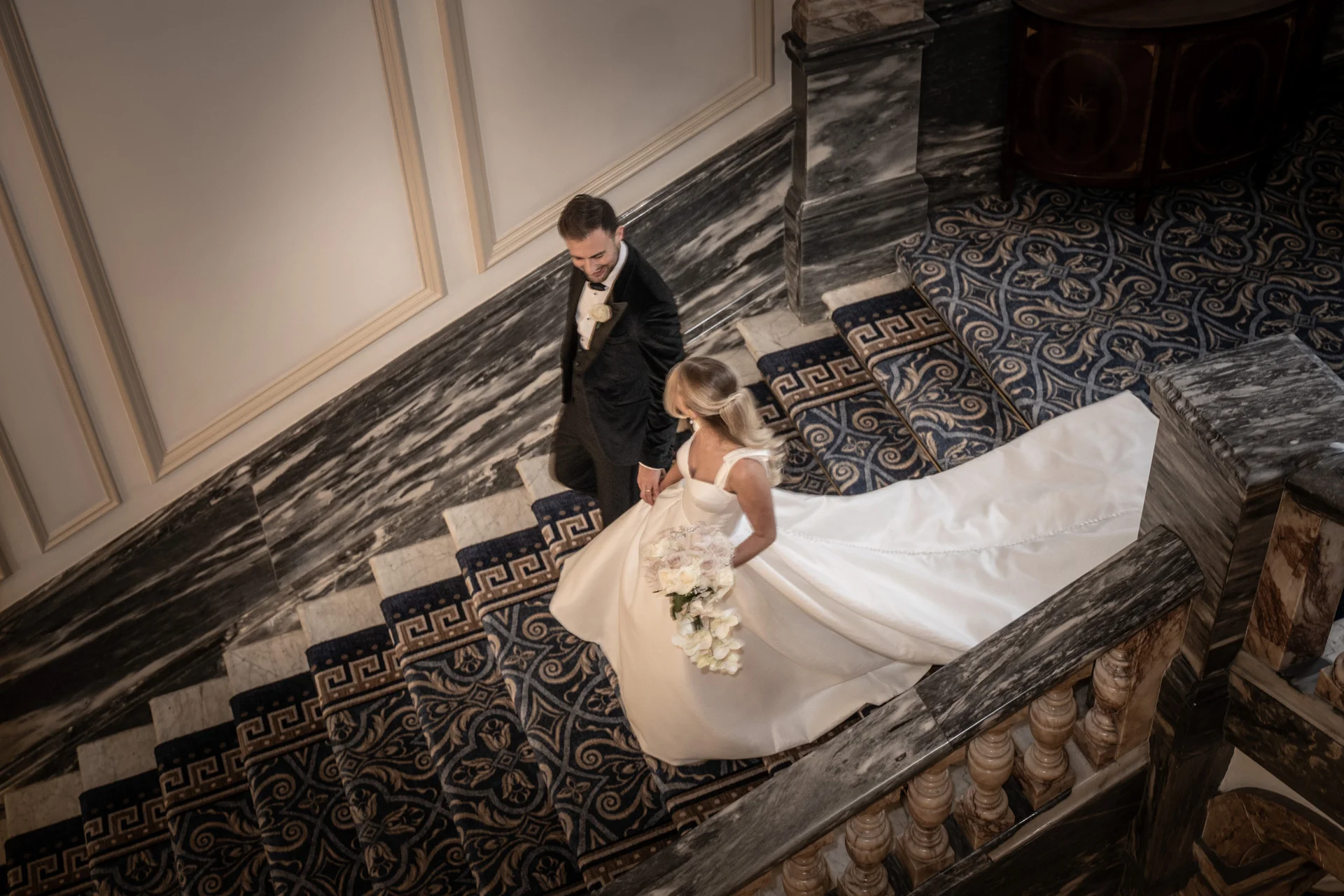 A newlywed couple descends a grand marble staircase, with the bride holding a cascading white floral bouquet and her long satin train trailing behind them.