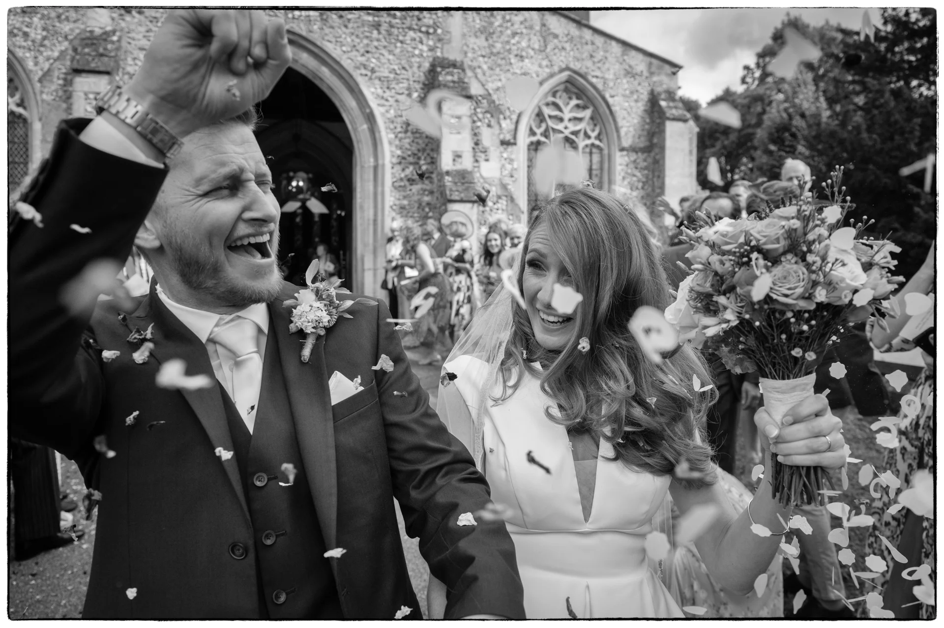A joyful newlywed couple laugh ecutatically as they walk out of a stone church amidst a flurry of falling confetti.