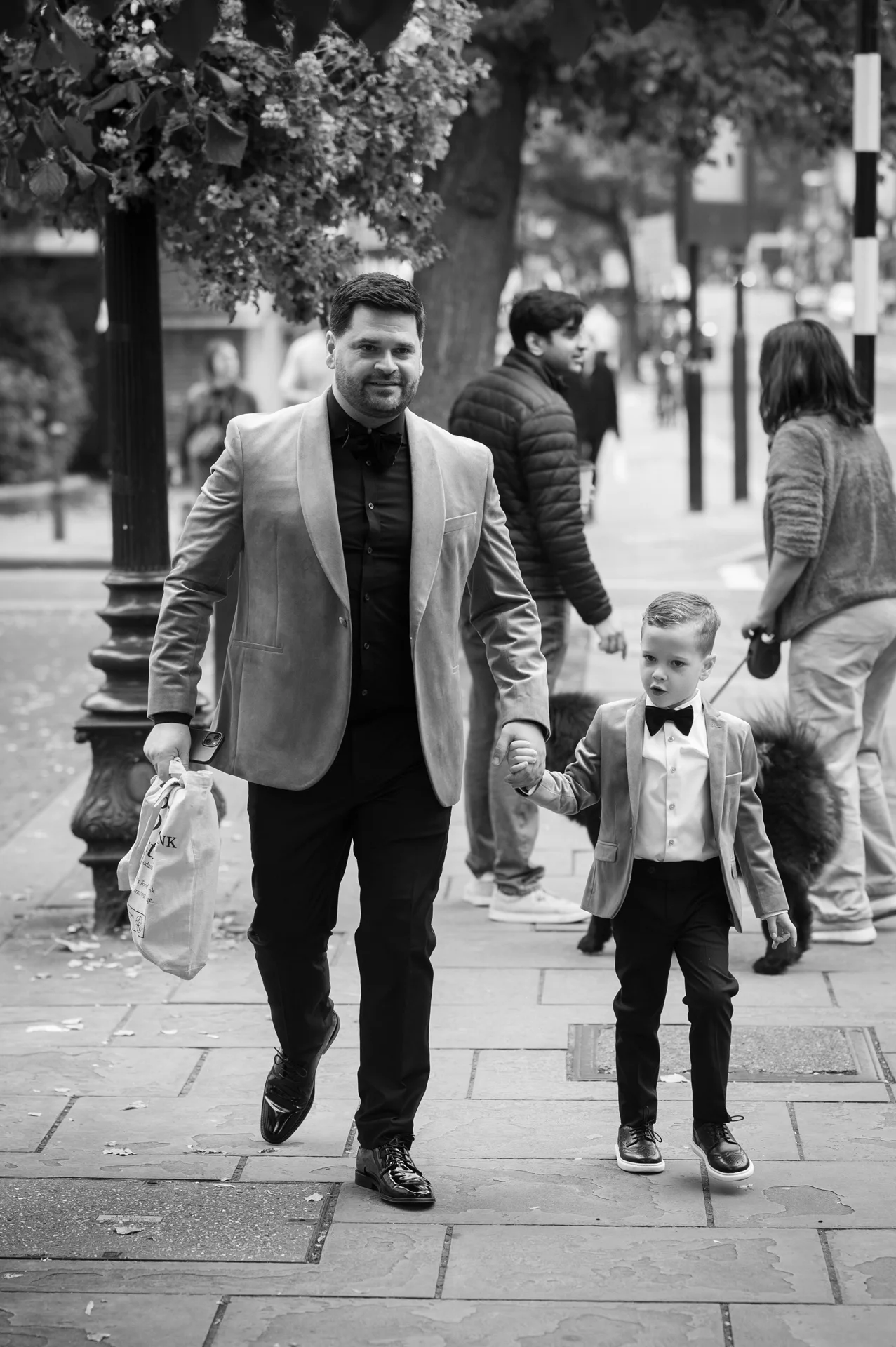 A groom and a page boy in matching velvet jackets and bow ties walk hand in hand down a city street.