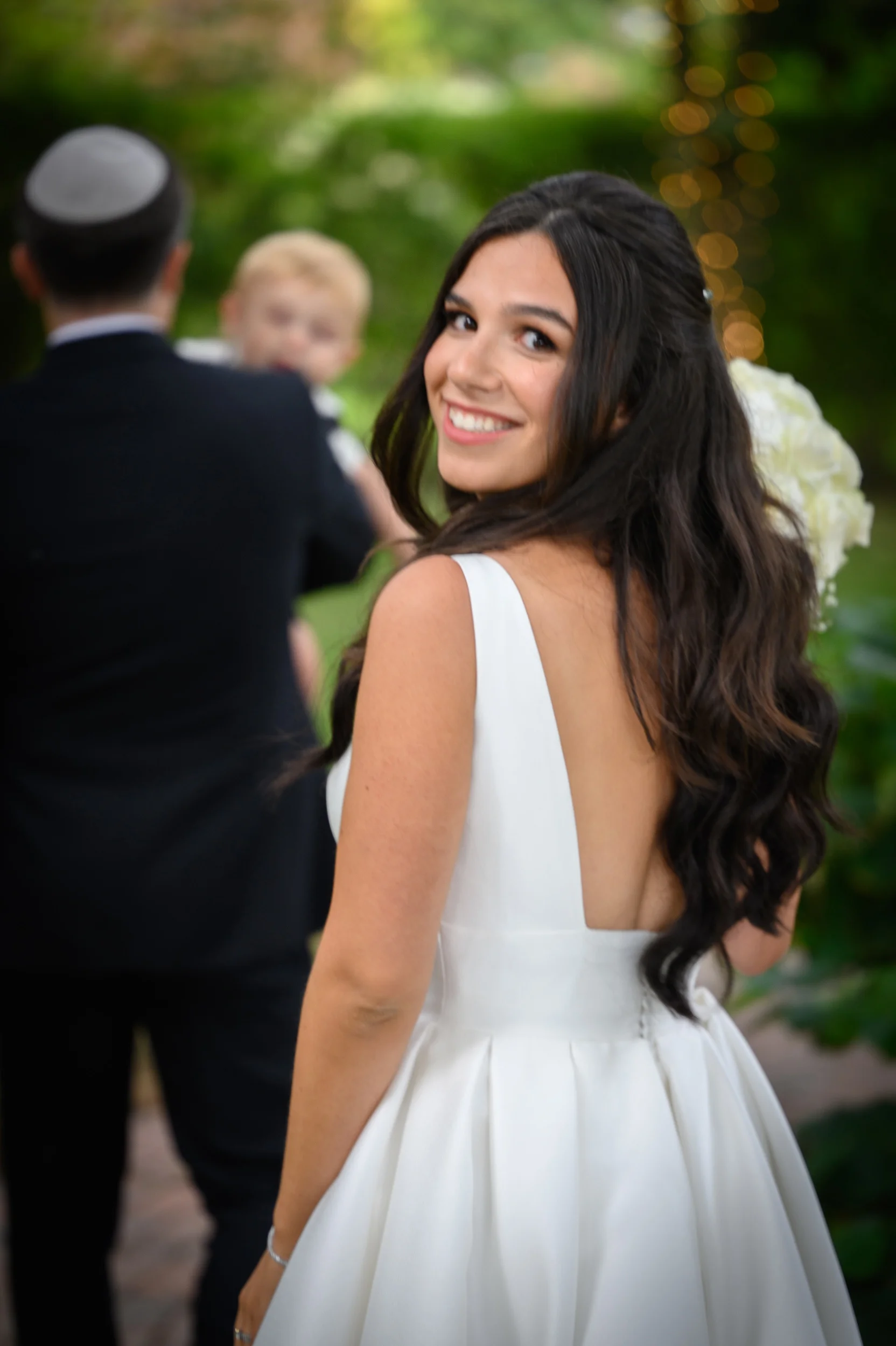 A smiling bride looks back over her shoulder wearing a modern white dress with a v-neckline and open back, her long dark hair styled in soft waves.