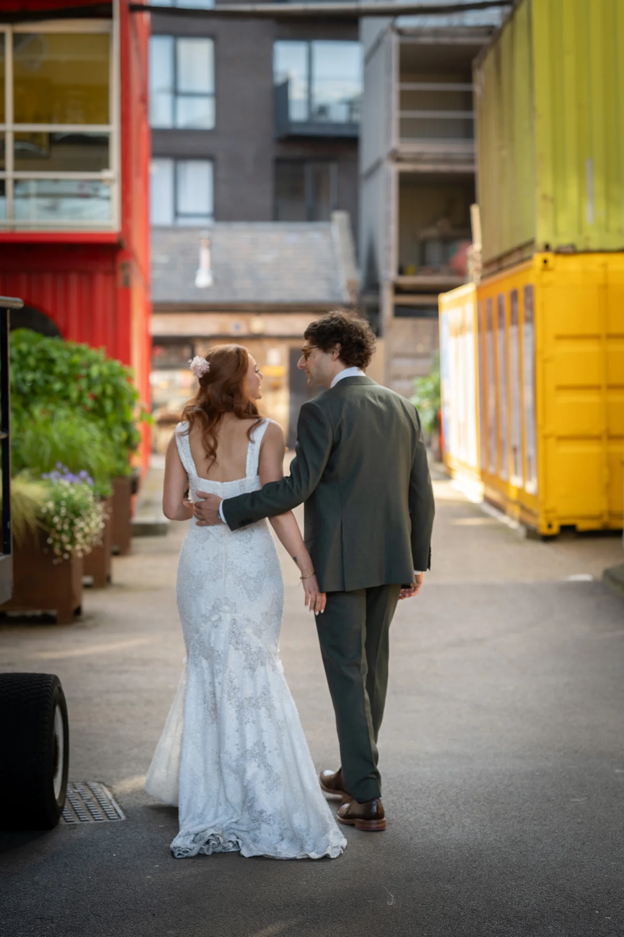 A bride and groom walk hand in hand down an urban alleyway framed by colourful shipping containers, sharing a sweet glance over their shoulders.