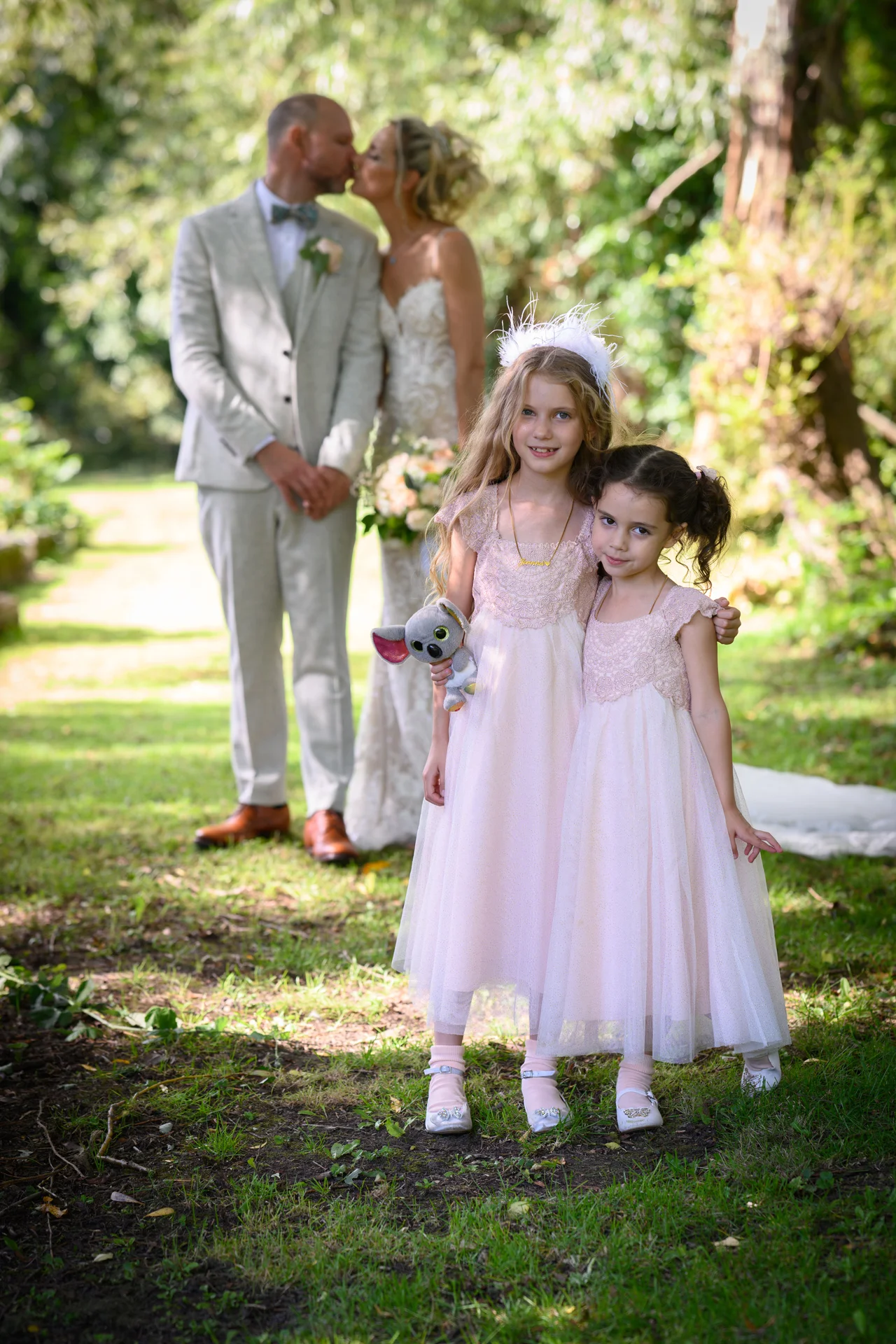 Two flower girls in matching pink dresses pose outdoors in a woodland setting while the bride and groom kiss softly in the softly blurred background.