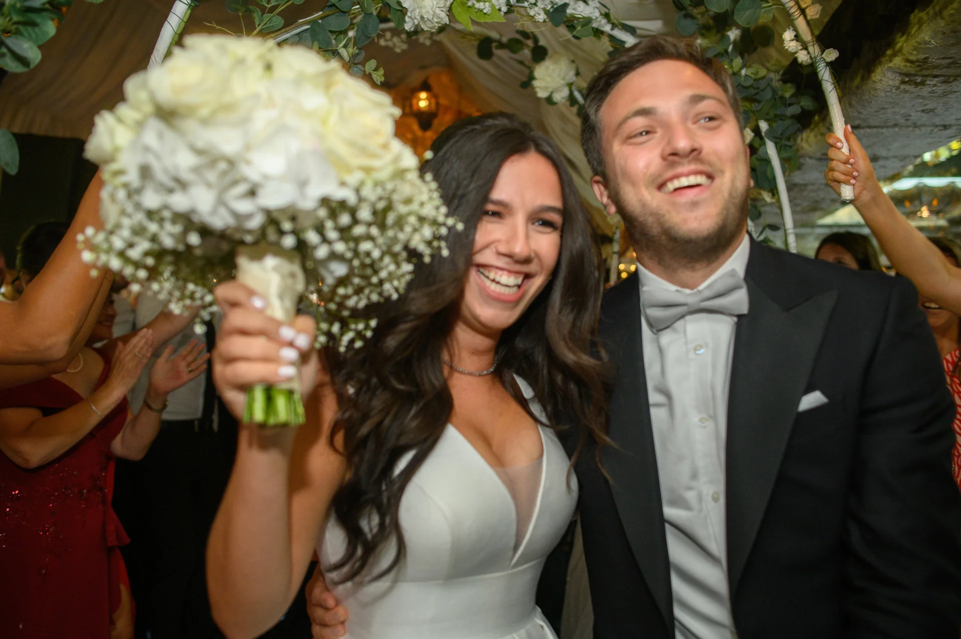 A joyful newlywed couple laughs happily as they walk through a flower-adorned archway, with the bride proudly holding up a white bouquet.