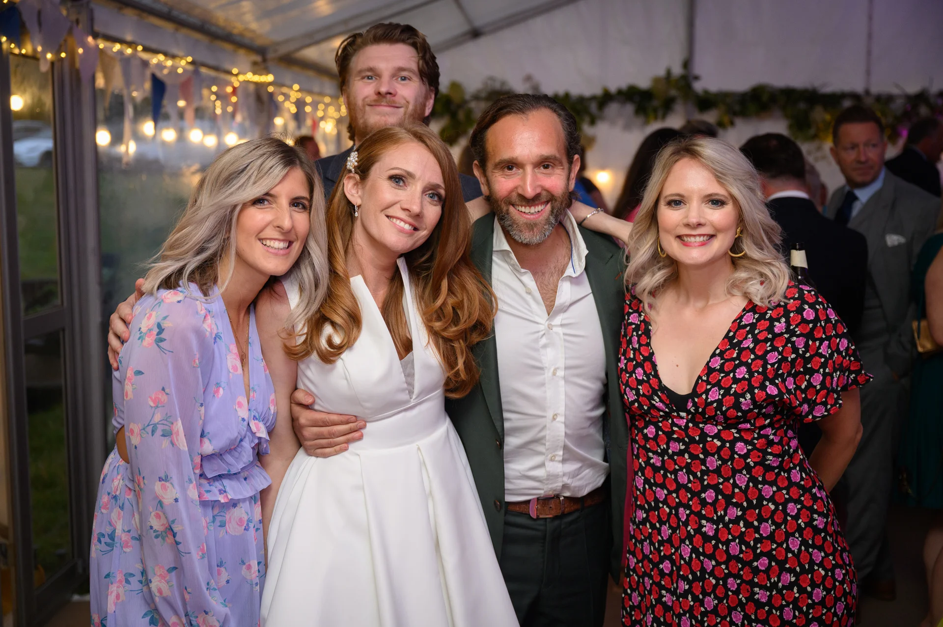 A newlywed couple and their friends pose and smile together inside a brightly lit marquee at a wedding evening reception.