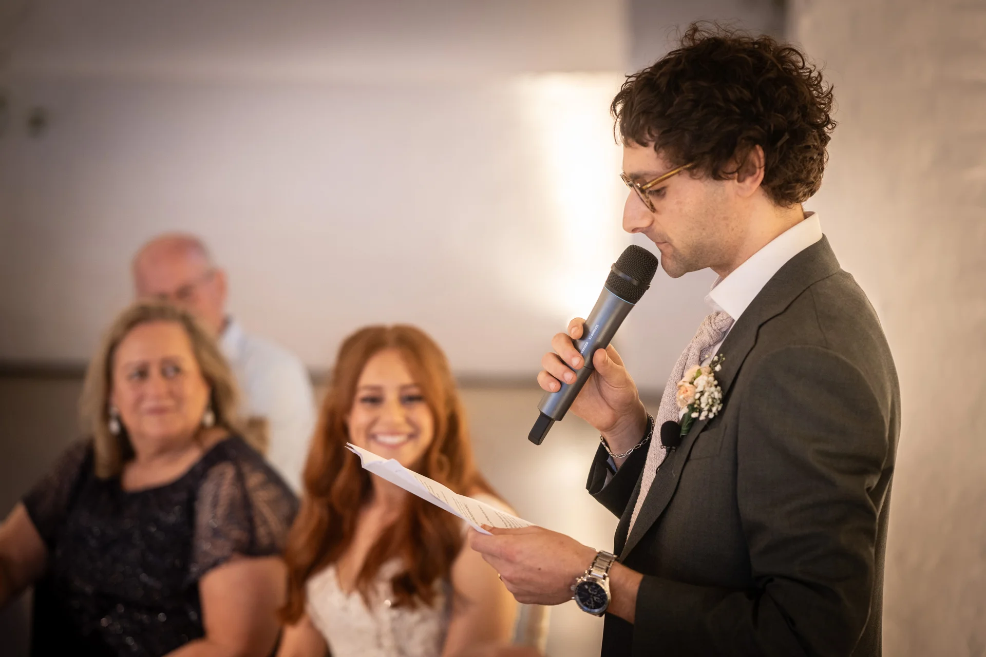 A groom in a grey suit delivers his wedding speech into a microphone while holding notes, with smiling guests seated in the background.