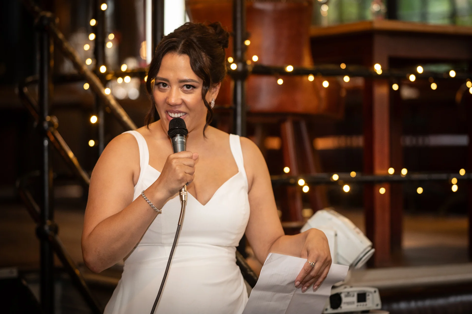 A smiling bride delivers her wedding speech into a microphone, holding notes in her other hand against a backdrop of glowing fairy lights.