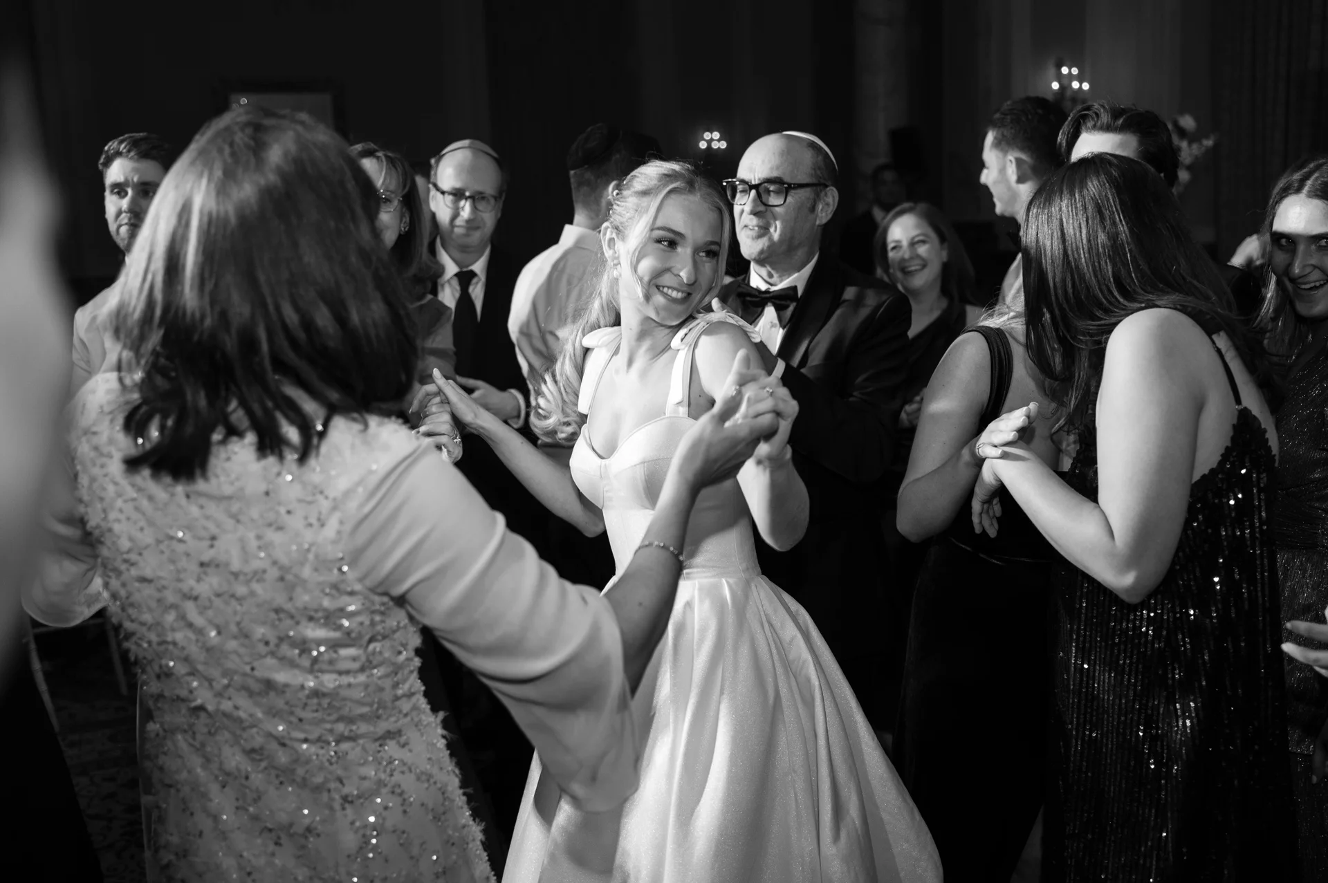 A smiling bride in a satin ball gown dances happily with guests in a lively black and white wedding reception scene.