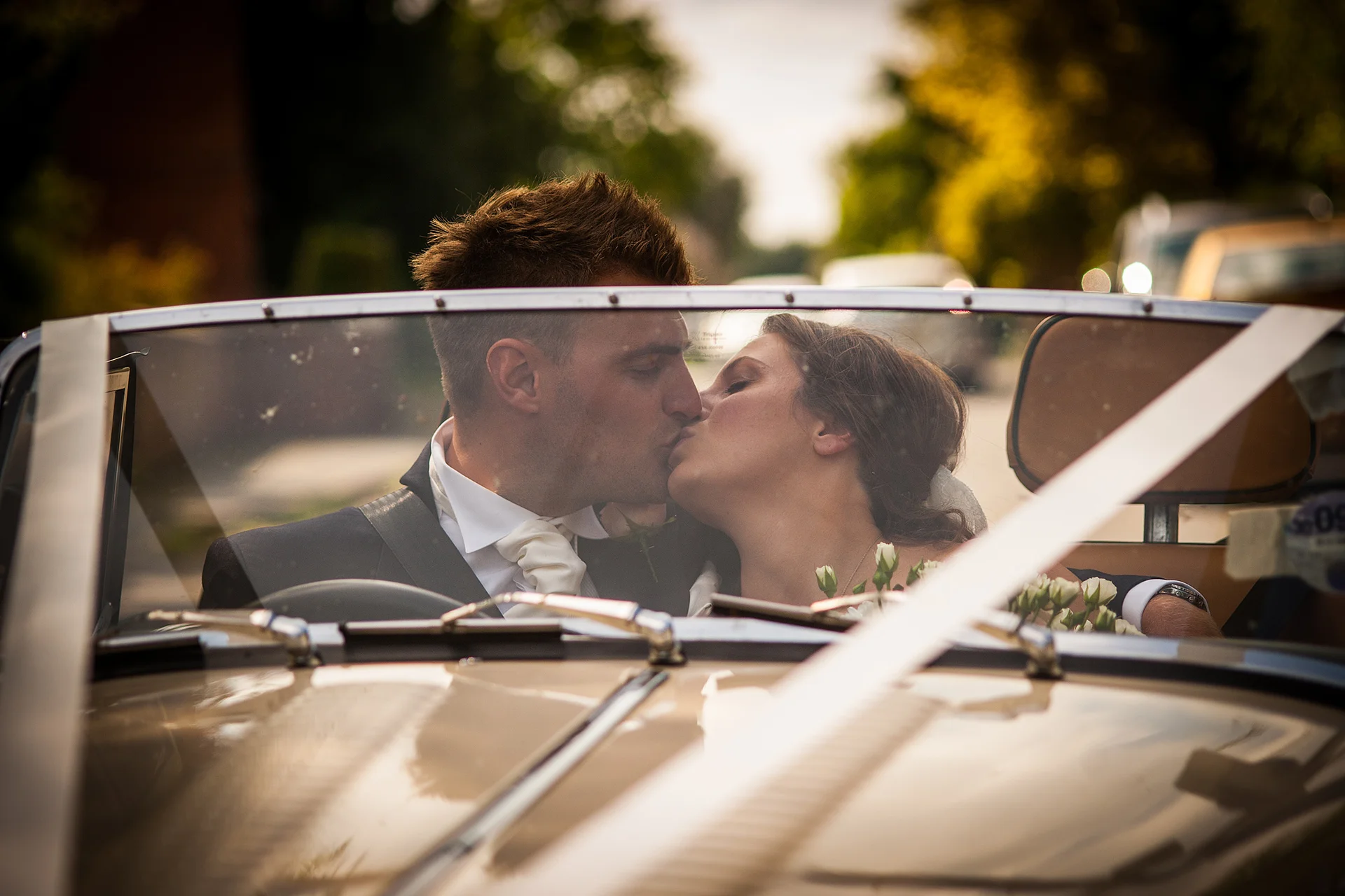 Newlywed bride and groom share a tender kiss inside a classic vintage wedding car decorated with ribbons and florals.