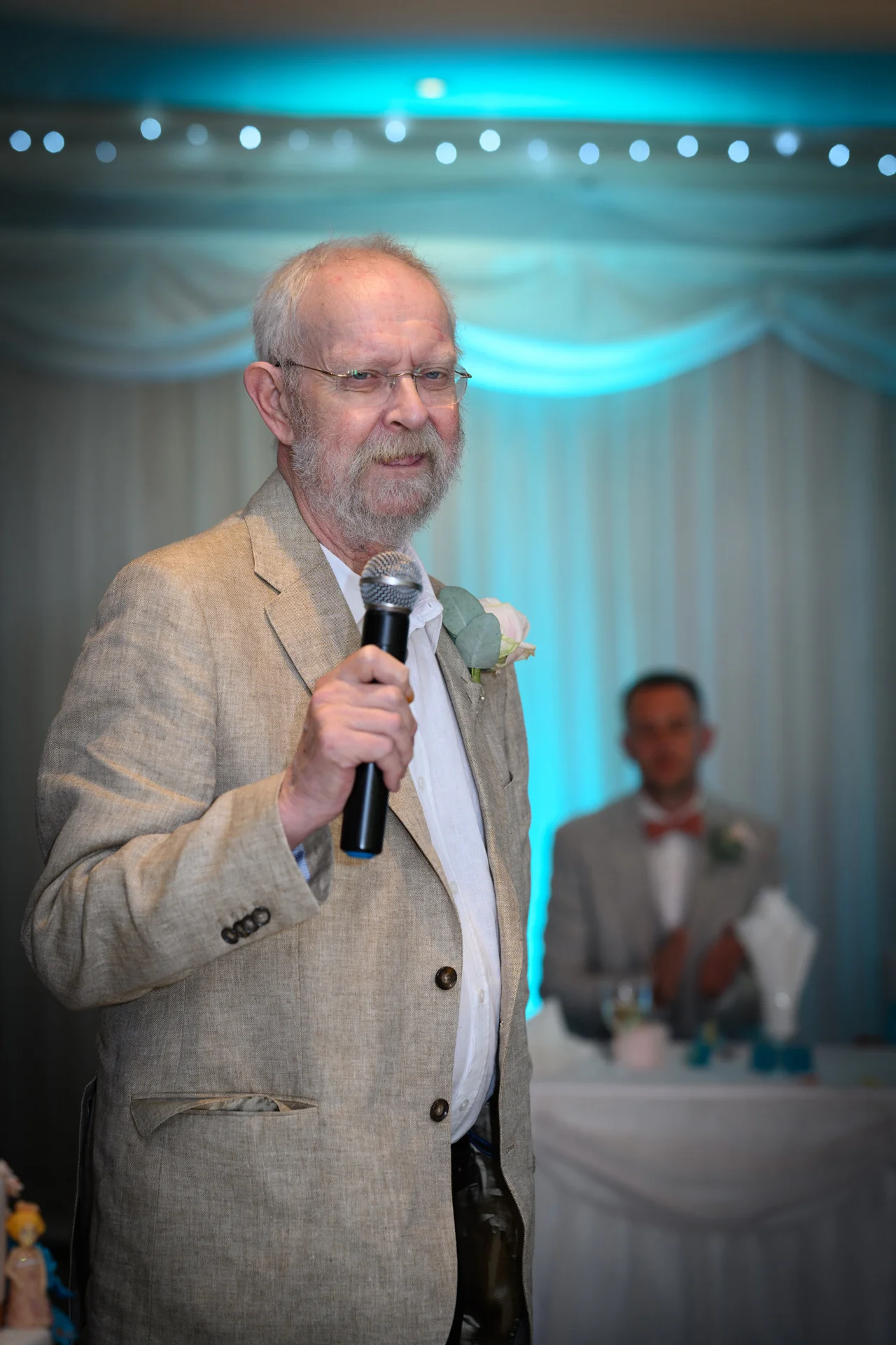 An older man wearing a linen jacket delivers a speech into a microphone during the wedding reception.