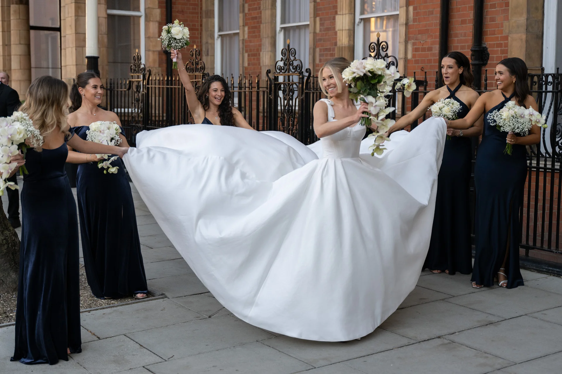A joyful bride in a voluminous white satin gown laughs as her bridesmaids hold out her dramatic skirt outside a red brick building with black iron railings.