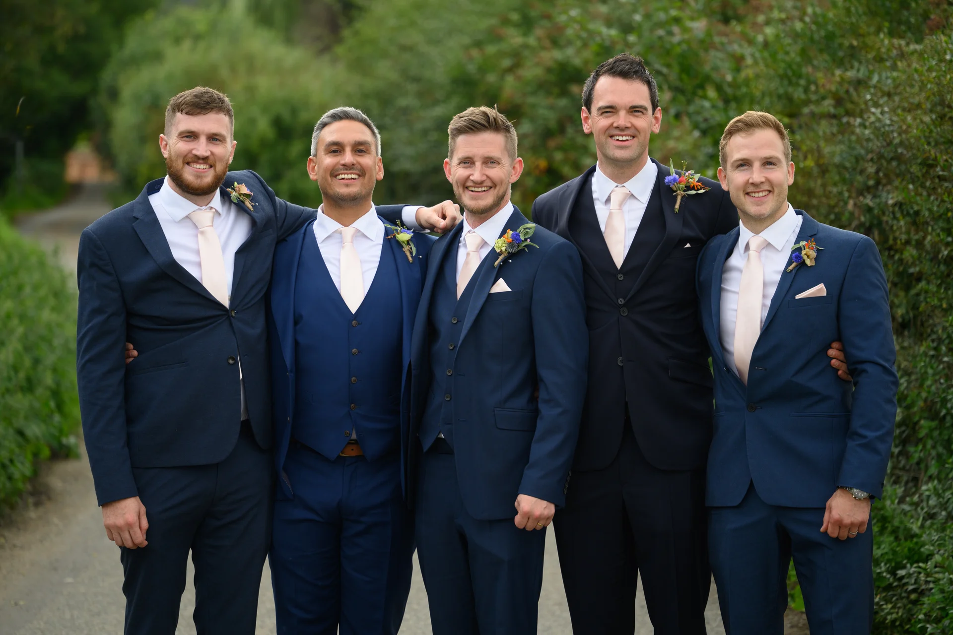 A group of smiling groomsmen in matching navy suits and pink ties pose together outdoors with floral buttonholes.