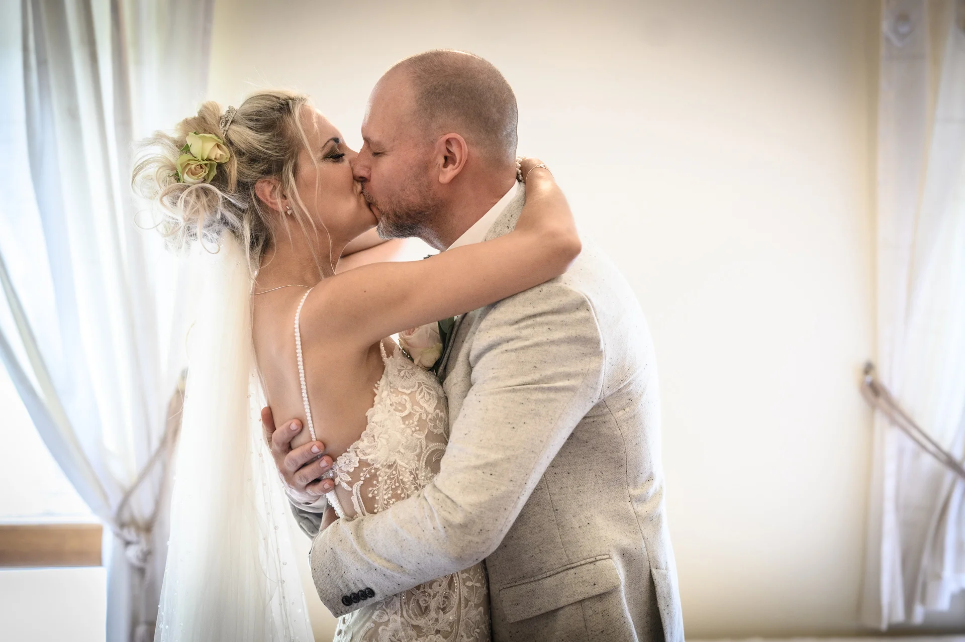 Newlyweds share an intimate first kiss indoors near a window, with the bride wearing a lace gown and an updo adorned with fresh flowers.