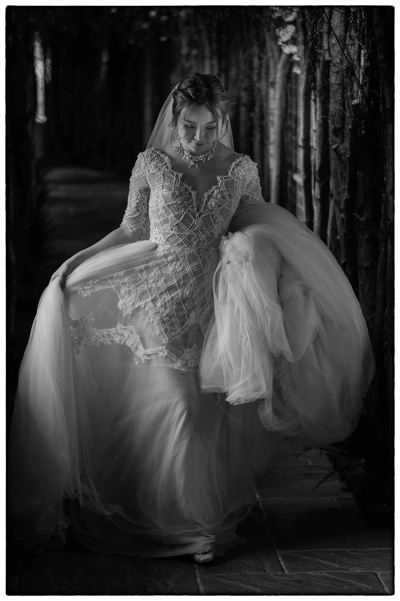 An east asian bride wearing a beaded v-neck gown and veil walks through a dimly lit woodland pathway while holding her voluminous tulle dress.