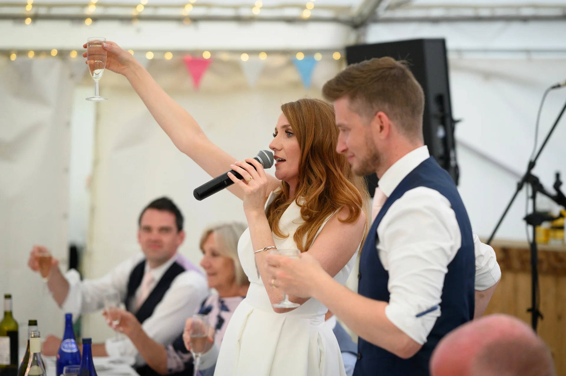 A bride with long red hair delivers a speech into a microphone inside a marquee while raising a glass of champagne, accompanied by her groom.