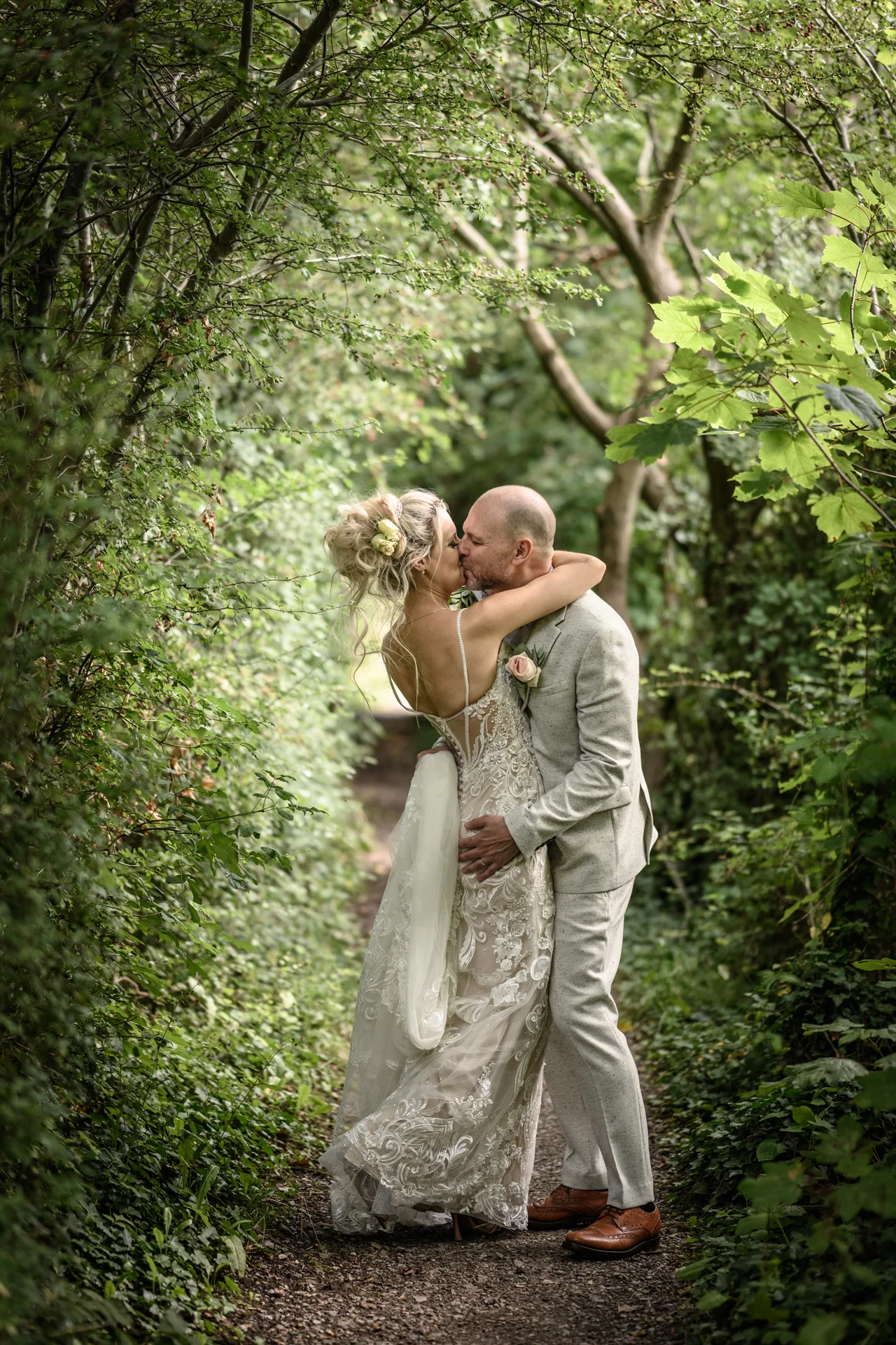 Newlyweds share a tender kiss along a lush, tree-lined path surrounded by vibrant green foliage in a woodland setting.