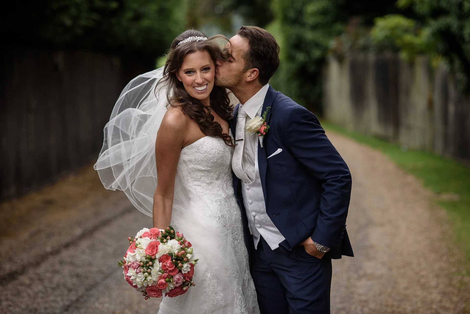 The groom lovingly kisses the smiling bride on the cheek outdoors while she holds a colourful pink and white rose bouquet, with her veil catching the breeze behind her.