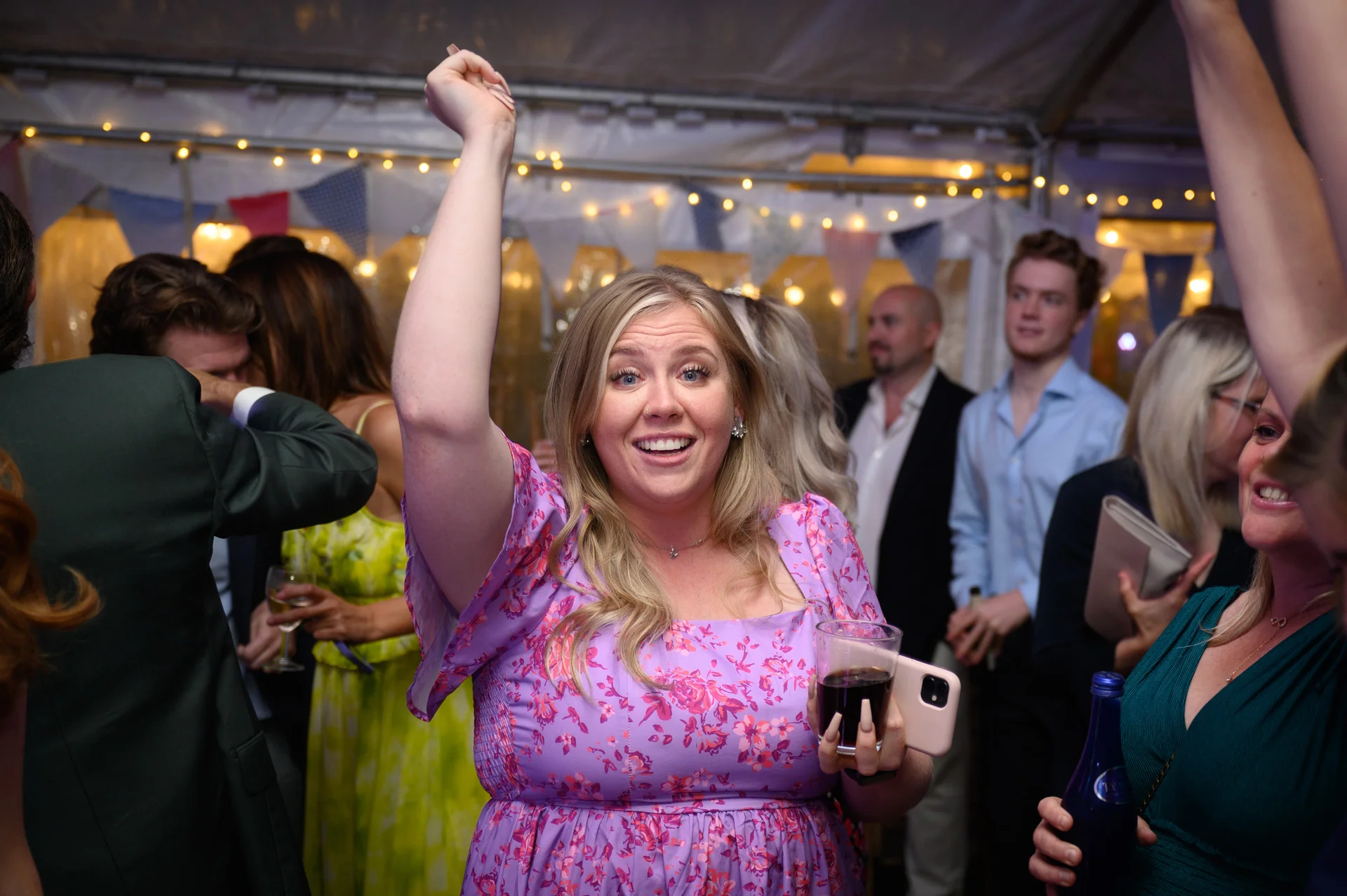 A joyful wedding guest in a floral print dress dances with her arm raised inside a marquee lit by festoon lights.